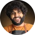 Smiling man with curly dark hair and beard wearing an orange shirt and a black apron against a brown background.
