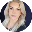 Portrait of a young woman with long blonde hair and light makeup, wearing a black top against a dark blue background.