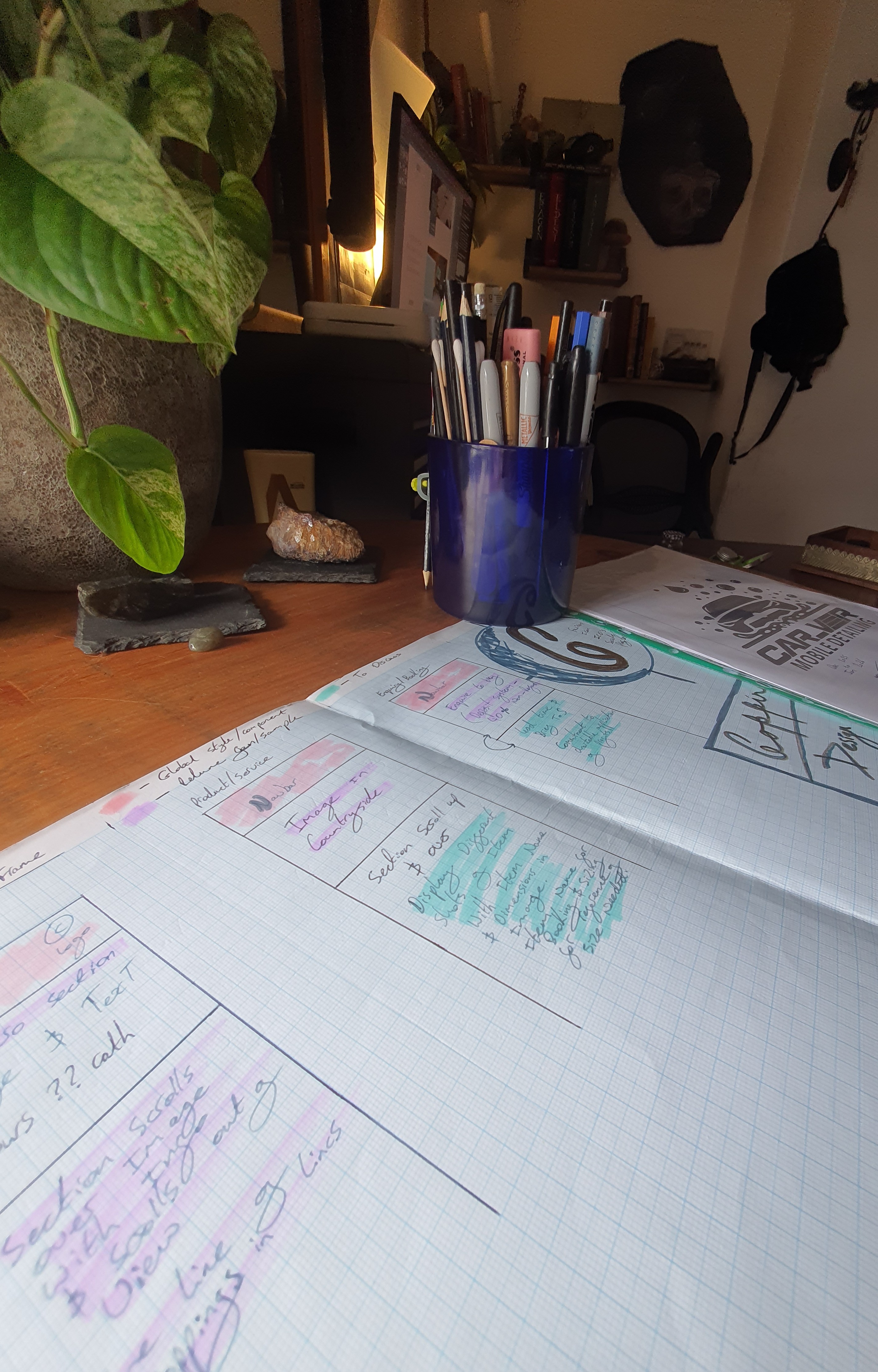 Close angle perspective of handwritten project notes and planning sketches resting on a wooden desk next to a dark travel mug and houseplant.