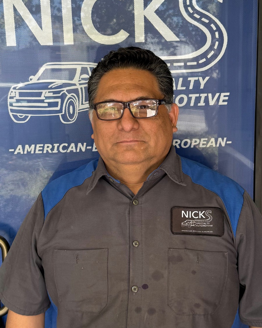Man wearing glasses and a gray uniform shirt with blue shoulder accents and a Nick's Specialty Automotive patch standing in front of a blue door with the Nick's Specialty Automotive logo and the words American-British-European.