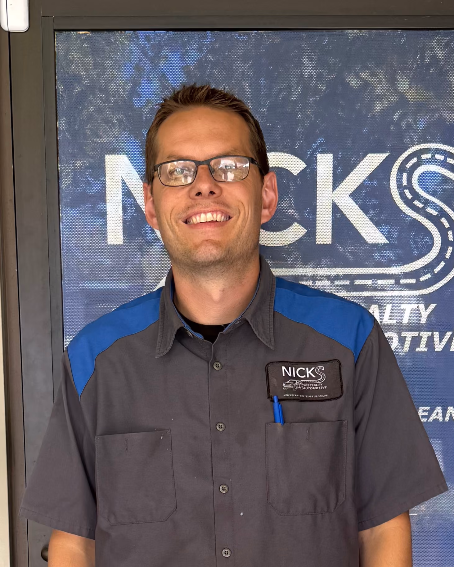 Smiling man wearing glasses and a grey work shirt with blue shoulder accents, standing in front of a window displaying a logo that reads 'NICKS Specialty Automotive'.