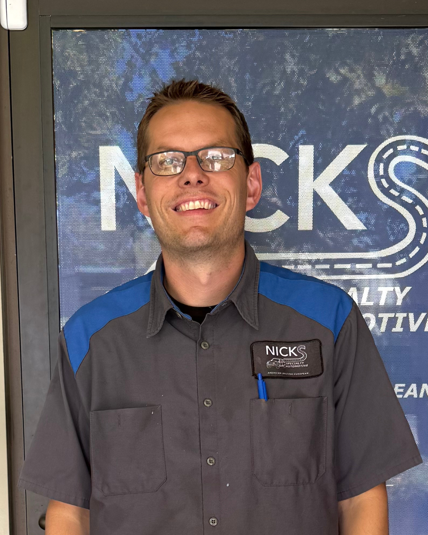 Smiling man wearing glasses and a grey work shirt with blue shoulder accents, standing in front of a window displaying a logo that reads 'NICKS Specialty Automotive'.