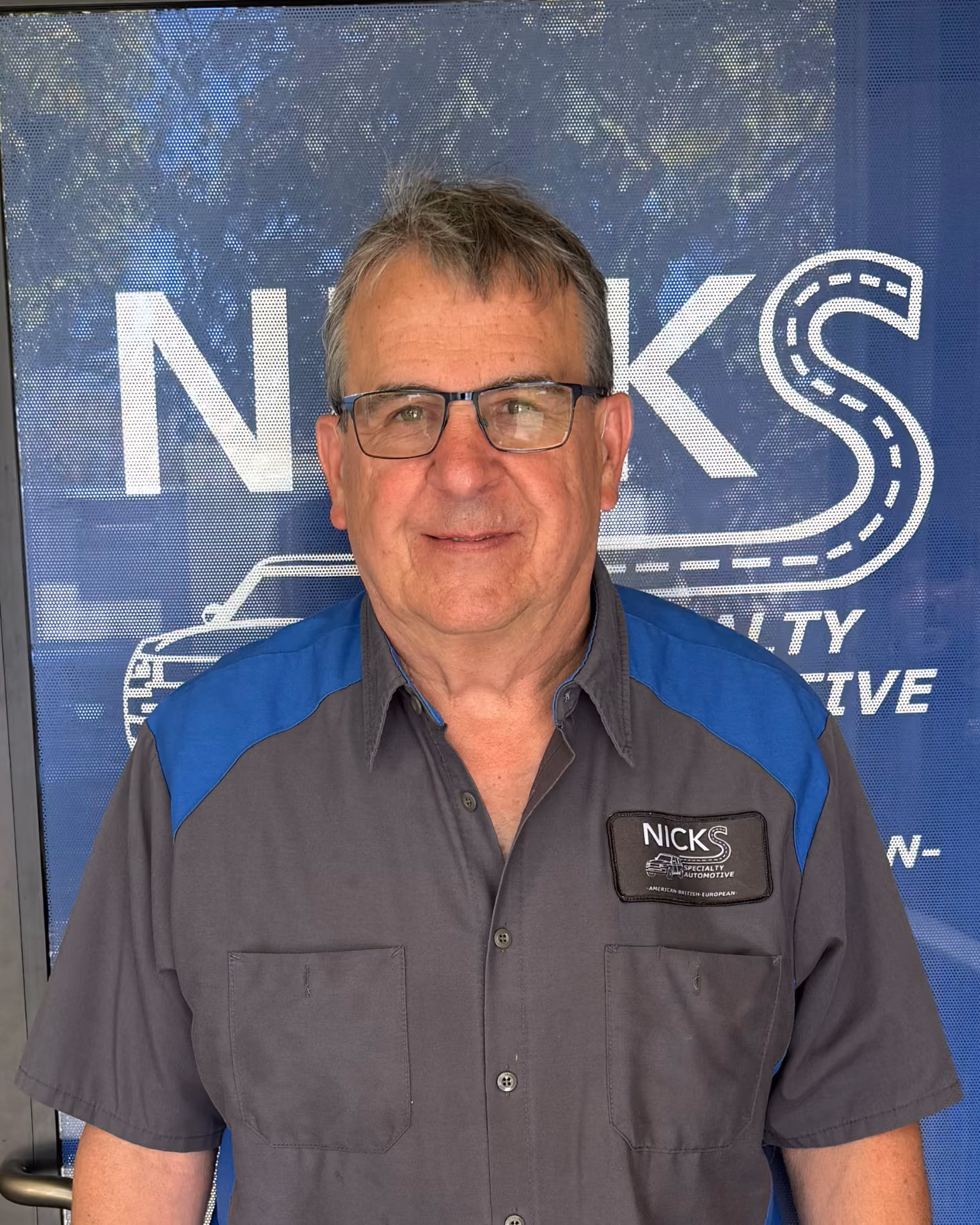 Middle-aged man with gray hair and glasses wearing a gray and blue work shirt with a 'Nicks Specialty Automotive' patch, standing in front of a blue backdrop with the 'Nicks' logo and a car graphic.