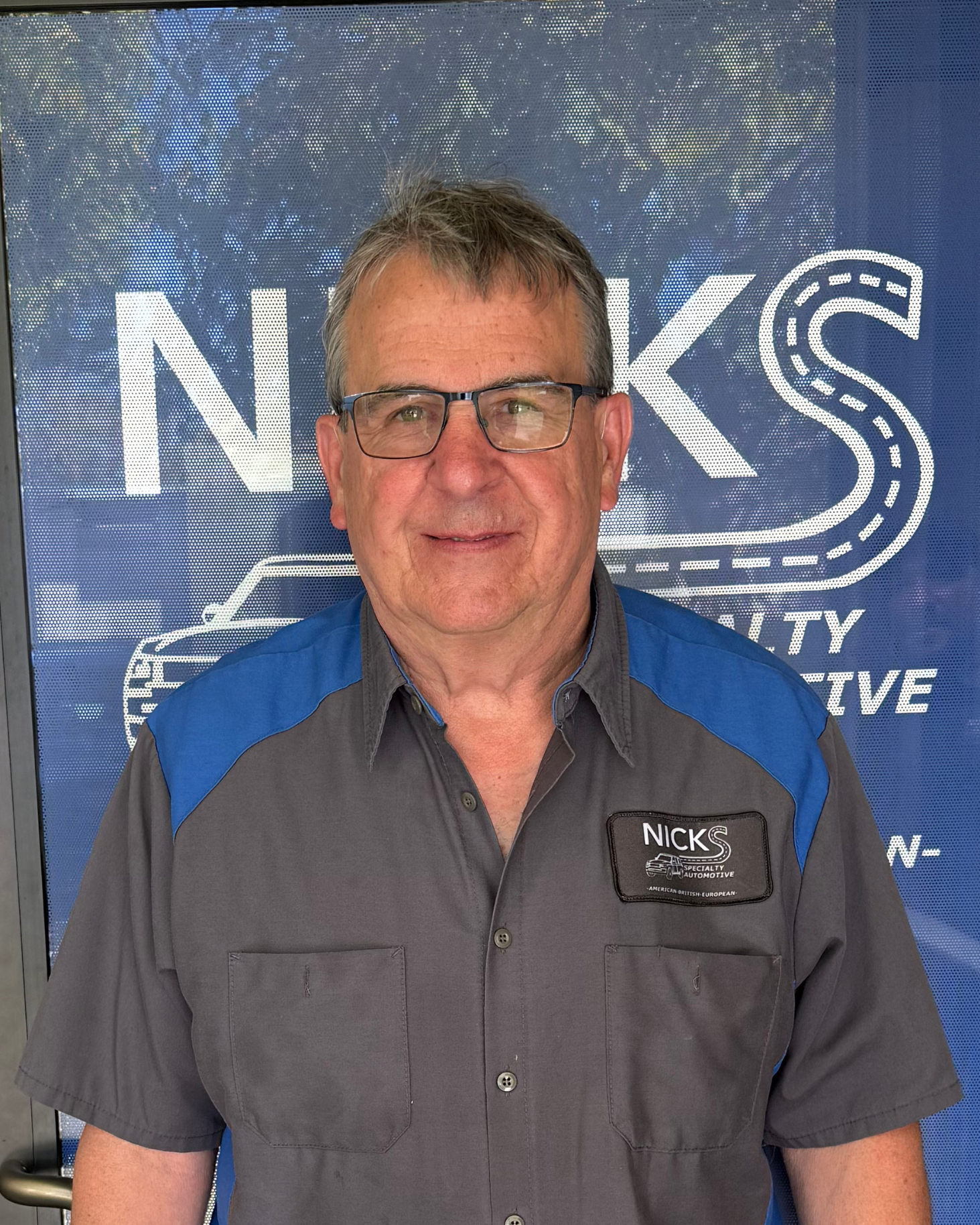 Middle-aged man with gray hair and glasses wearing a gray and blue work shirt with a 'Nicks Specialty Automotive' patch, standing in front of a blue backdrop with the 'Nicks' logo and a car graphic.