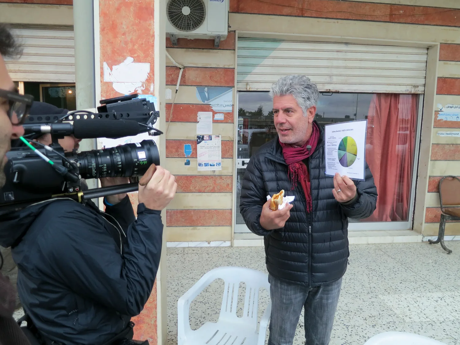 Anthony Bourdain eats street food while being filmed outside a shop in Libya.
