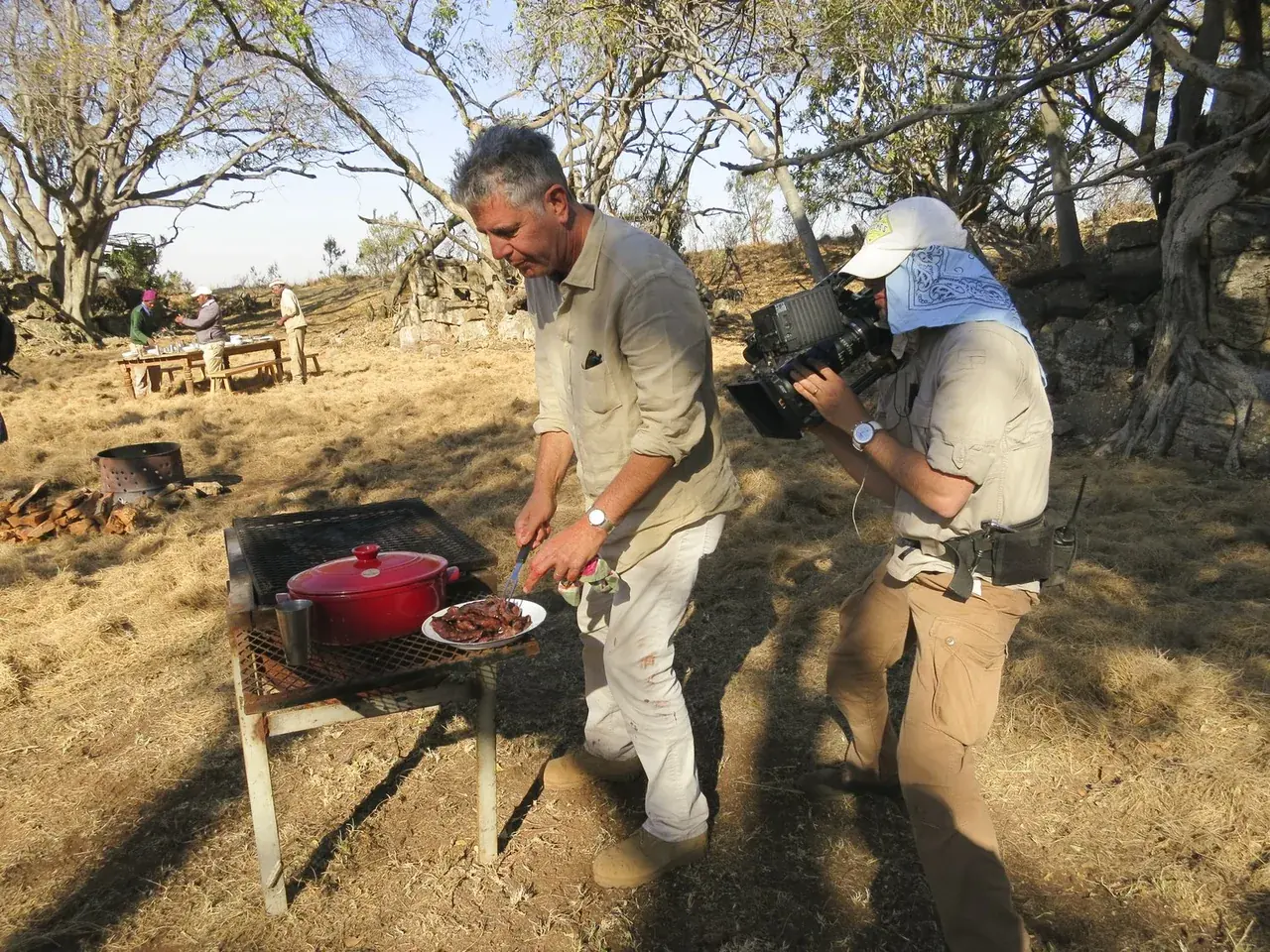 Anthony Bourdain plates food beside a barbecue as a camera operator films him.