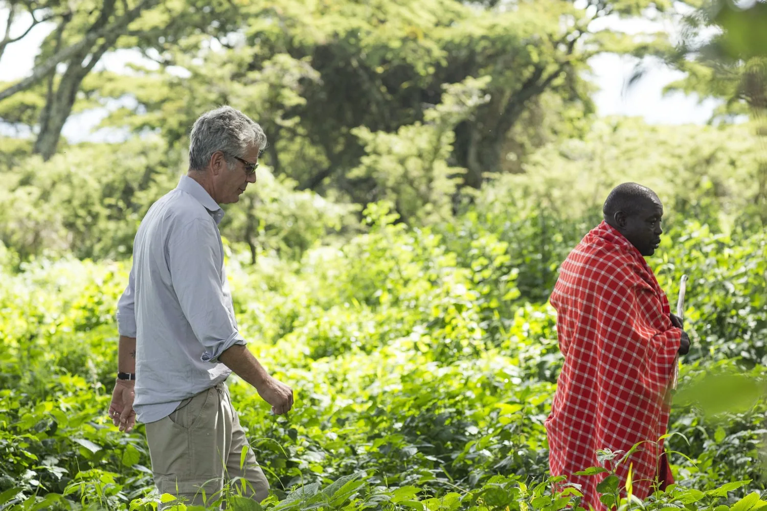 Anthony Bourdain walks through greenery beside a man wearing a red checked wrap.