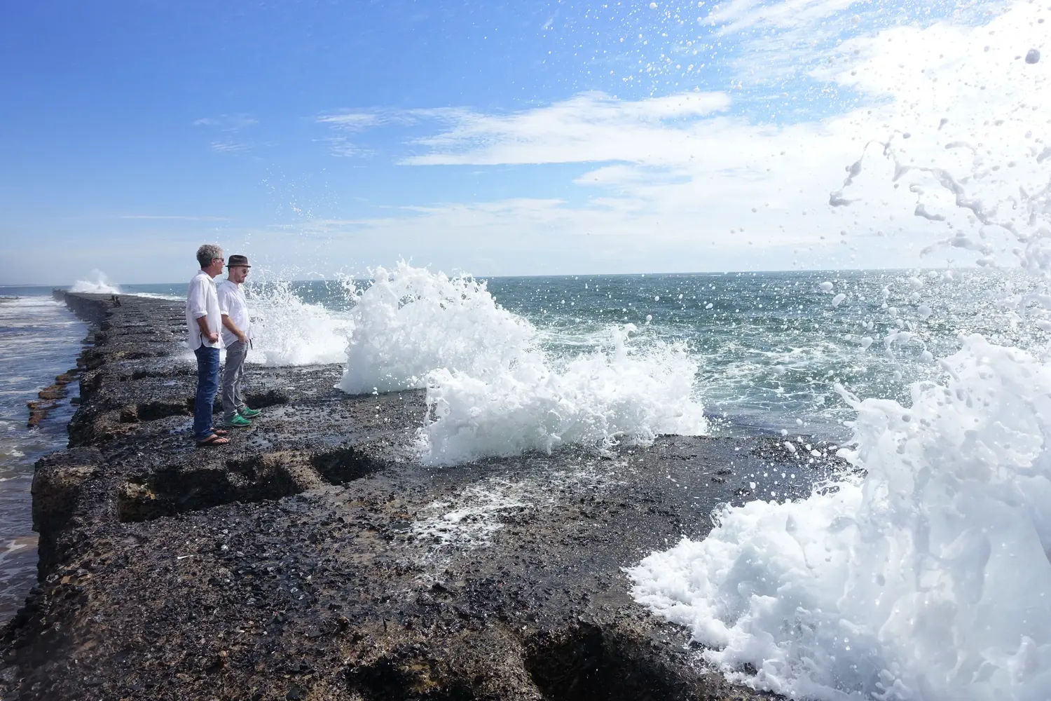 Anthony Bourdain and Darren Aronofsky stand on a sea wall in Madagascar as a wave breaks beside them.
