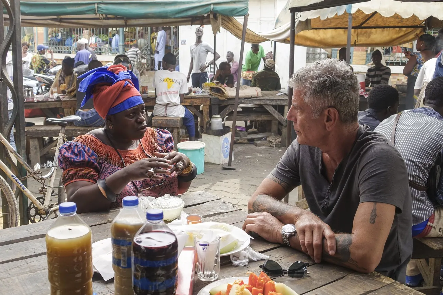 Anthony Bourdain talks with a woman over food at an outdoor market table in Senegal.
