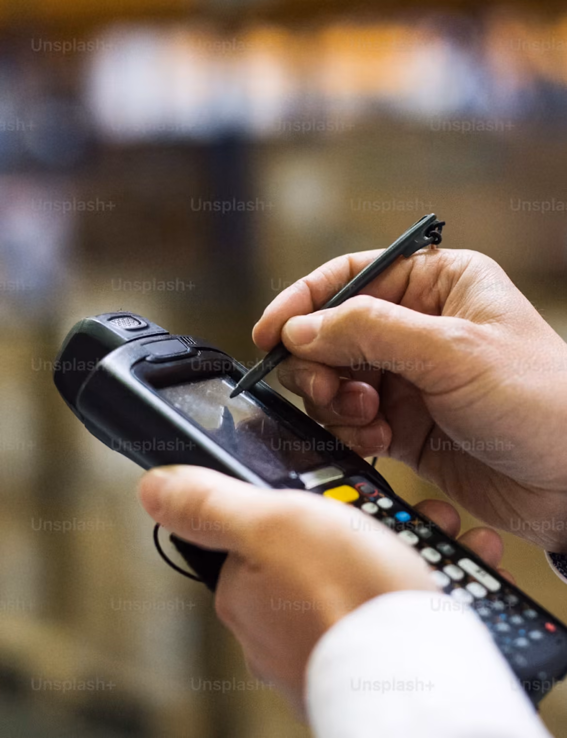 Eine Person bedient ein mobiles Handgerät mit einem Stift, das wie ein Scanner oder Terminal aussieht, vor unscharfem Hintergrund.