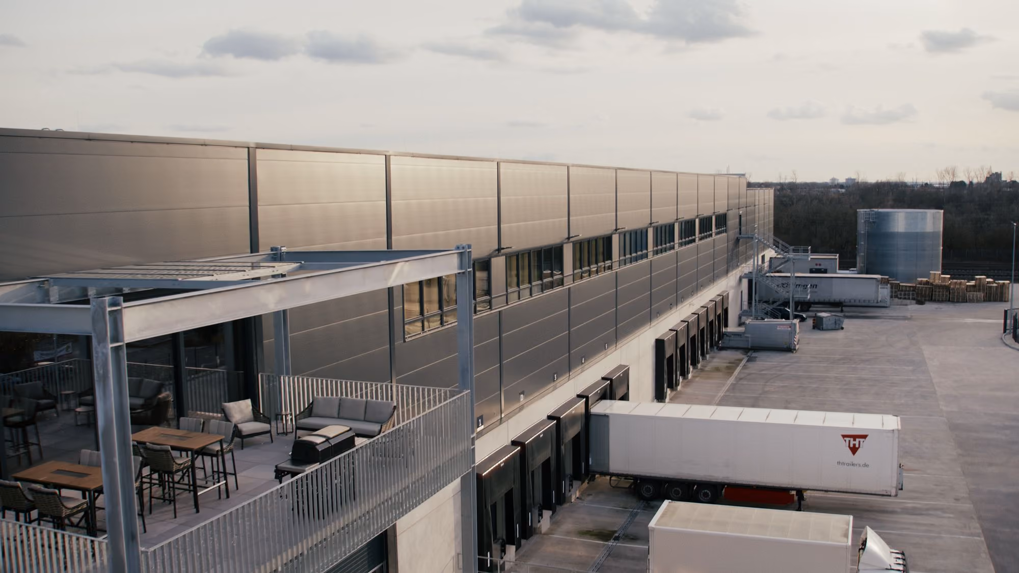 Außenansicht eines modernen Logistikzentrums mit einer Serie von Ladebuchten, an denen Lastwagen an- und abkuppeln, daneben Sitzbereiche mit Tischen und Stühlen auf einem Balkon.
