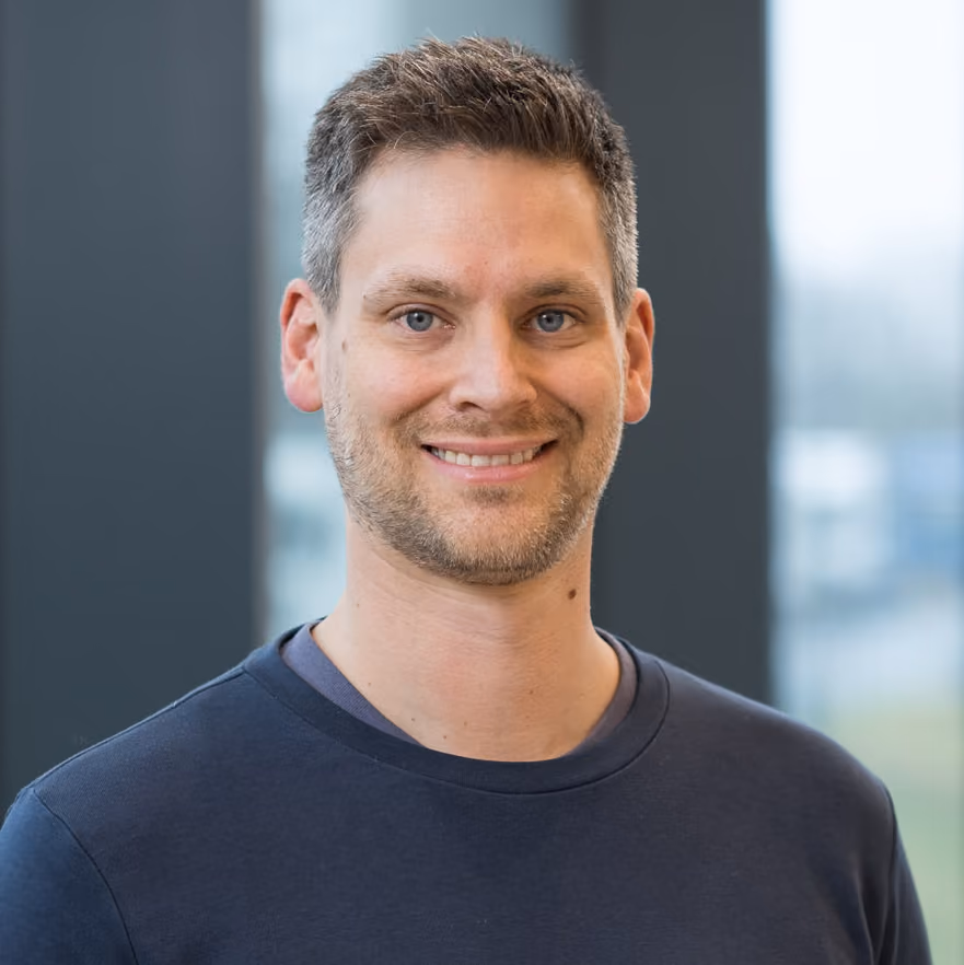 Porträt eines lächelnden Mannes mit kurzen braunen Haaren und Bart, tragen ein dunkelblaues T-Shirt, unscharfer Hintergrund mit großen Fenstern.