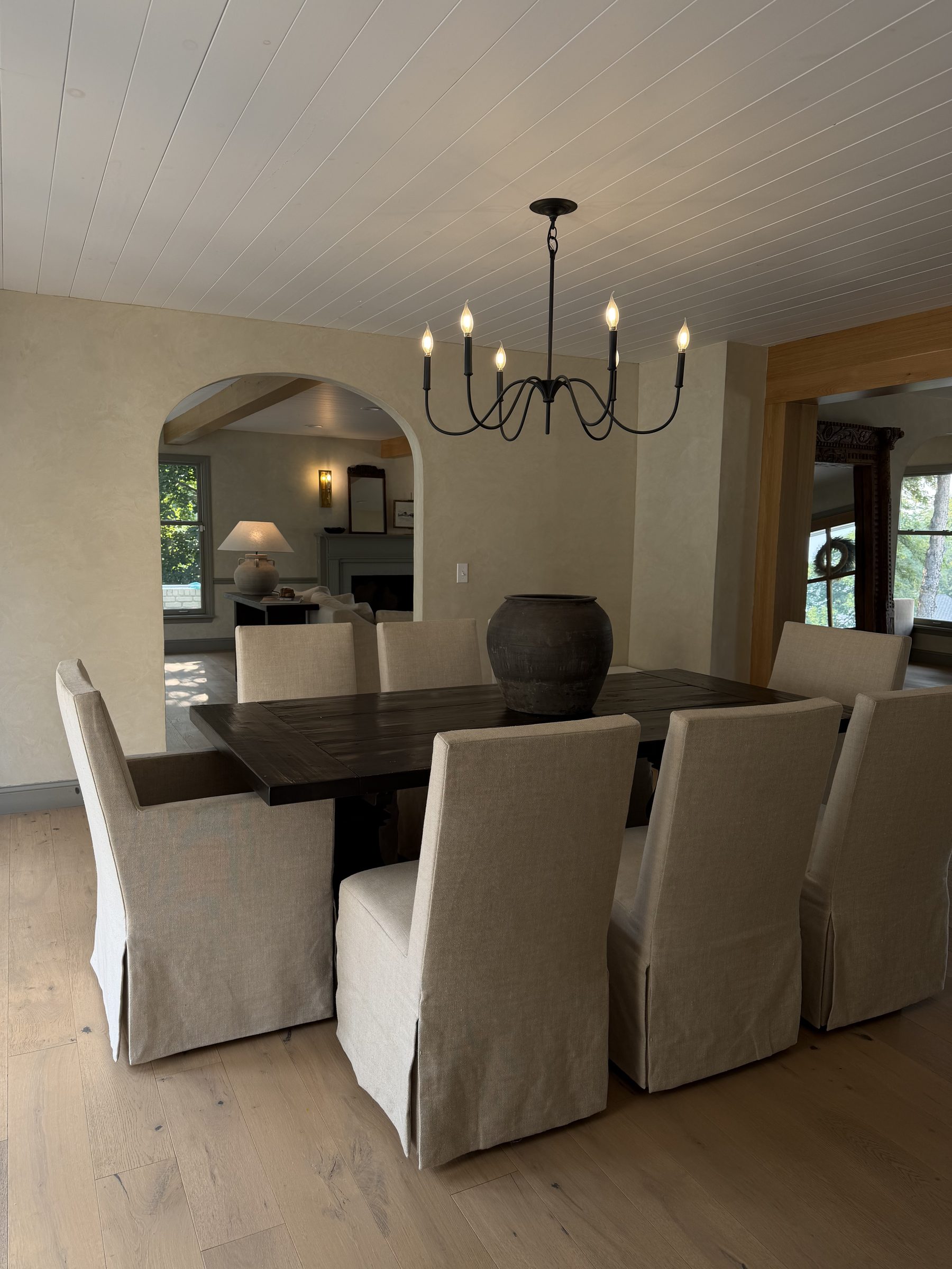 Dining room with a dark wood table, linen slipcovered chairs, iron candle chandelier, and an arched opening to the living room