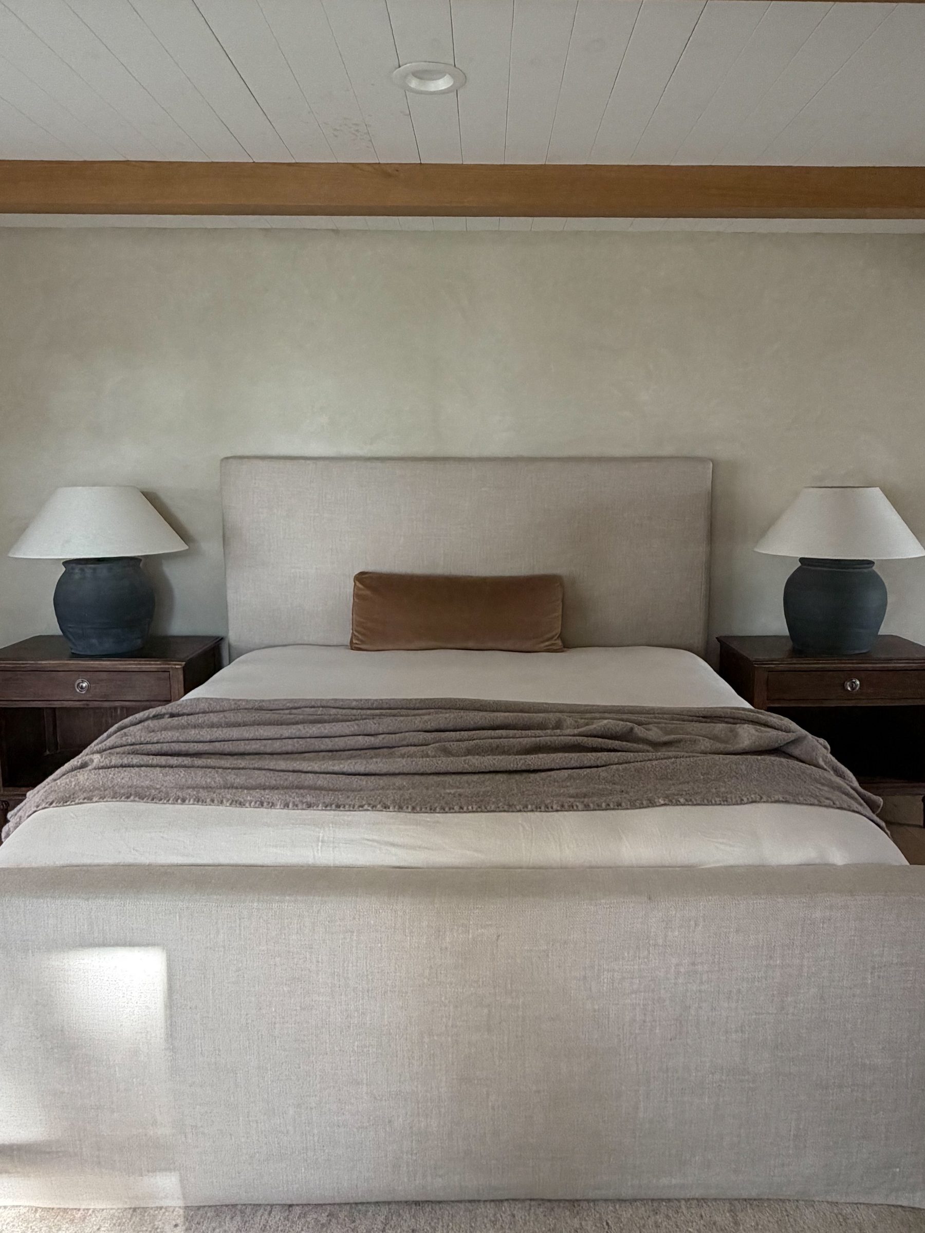 Primary bedroom with a linen upholstered bed, layered neutral bedding, plaster walls, and a pair of ceramic table lamps