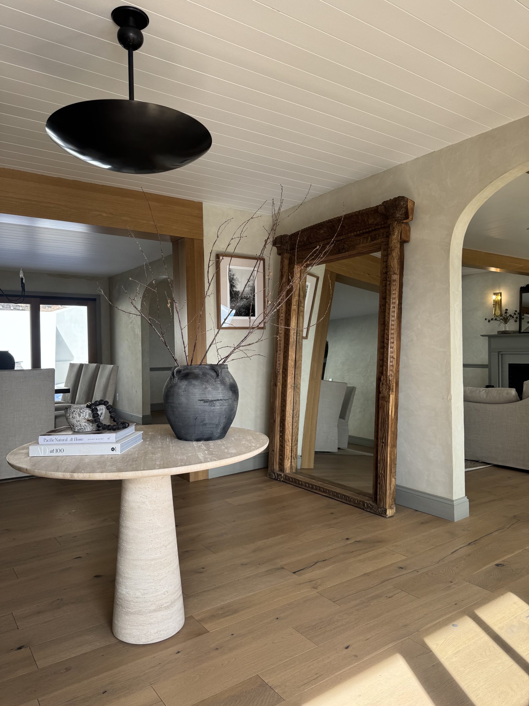 Entry foyer with a round travertine table, large antique carved mirror, and white oak floors, with sightlines into the dining and living rooms
