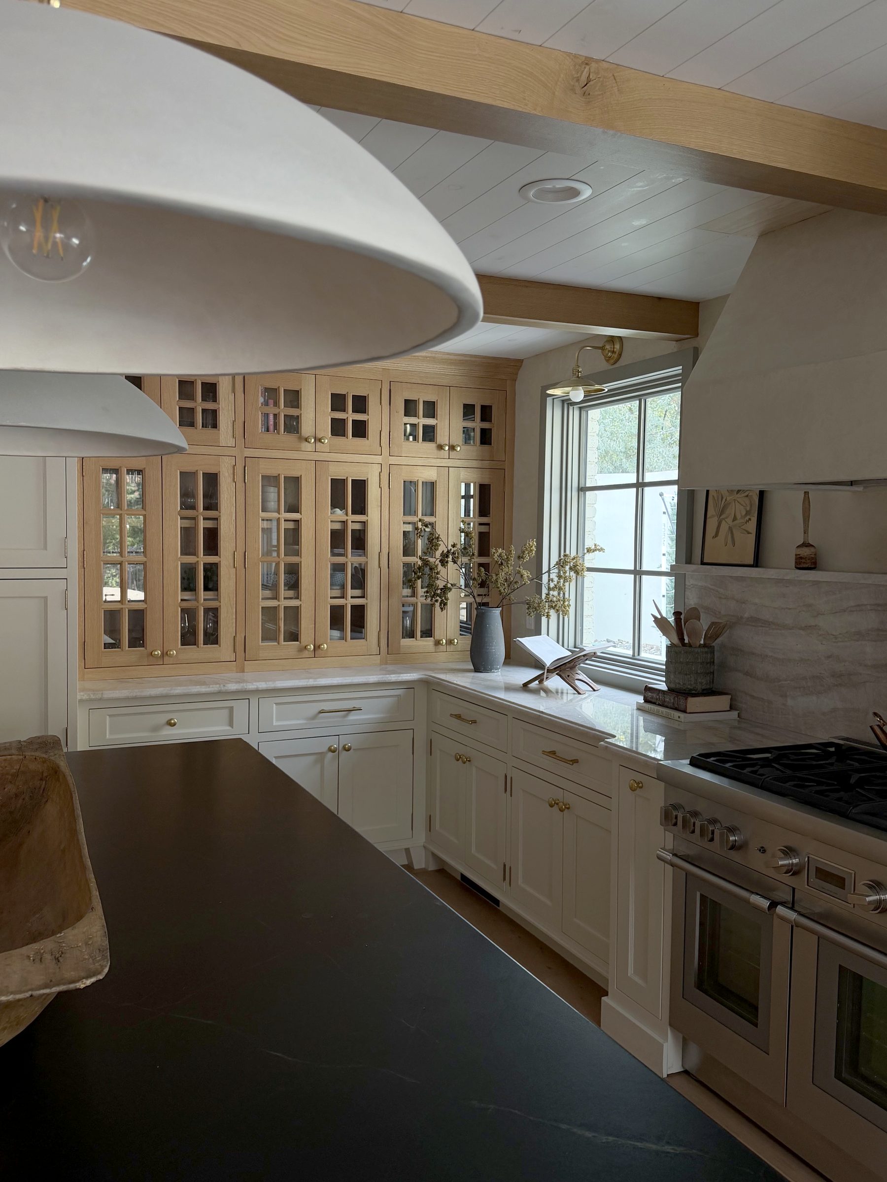 Kitchen with white oak glass-front cabinetry, Taj Mahal quartzite counters, brass hardware, a plaster range hood, and a soapstone island