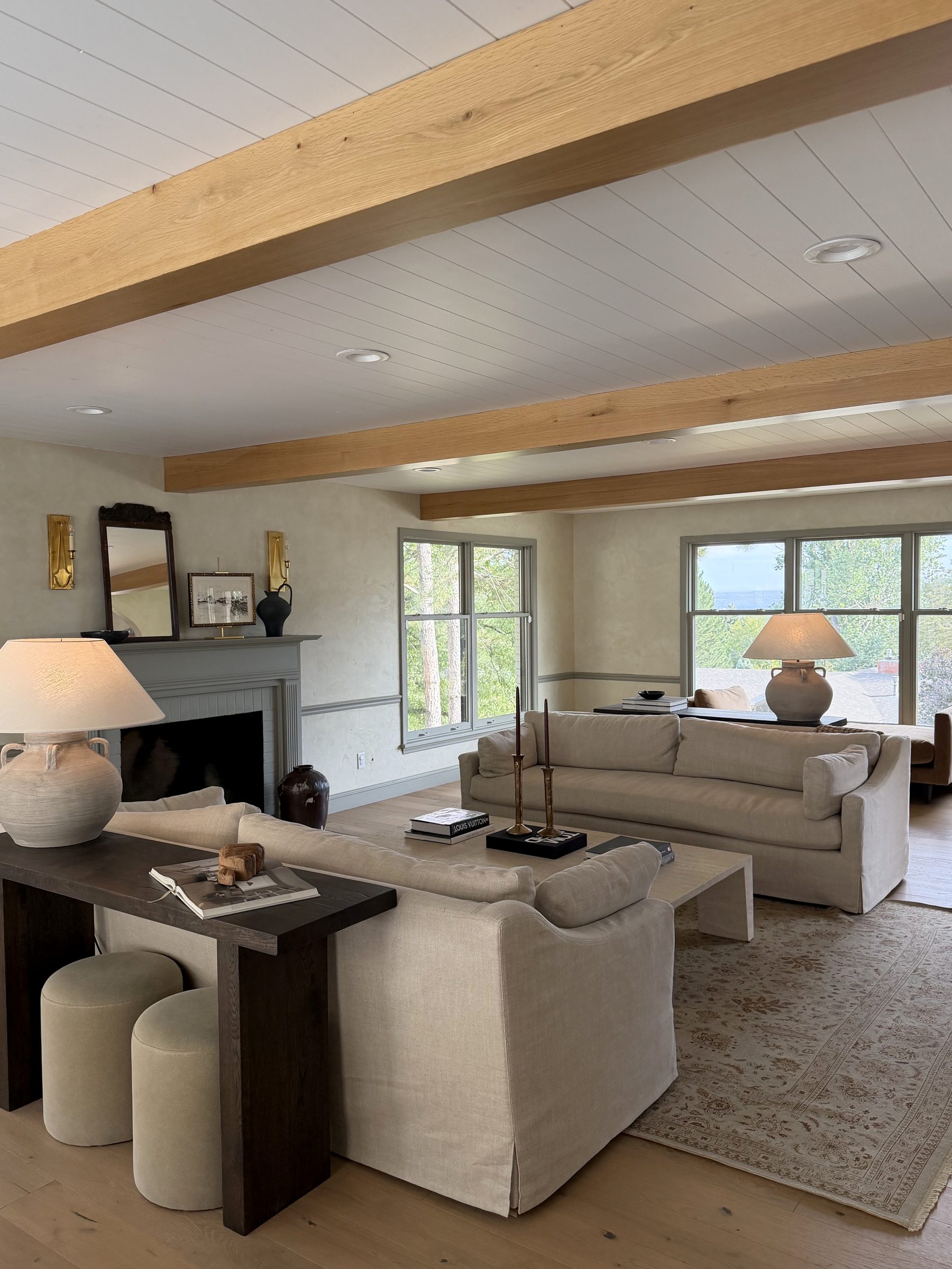 Living room with white oak ceiling beams, two linen sofas, a fireplace with brass sconces, and windows on multiple walls
