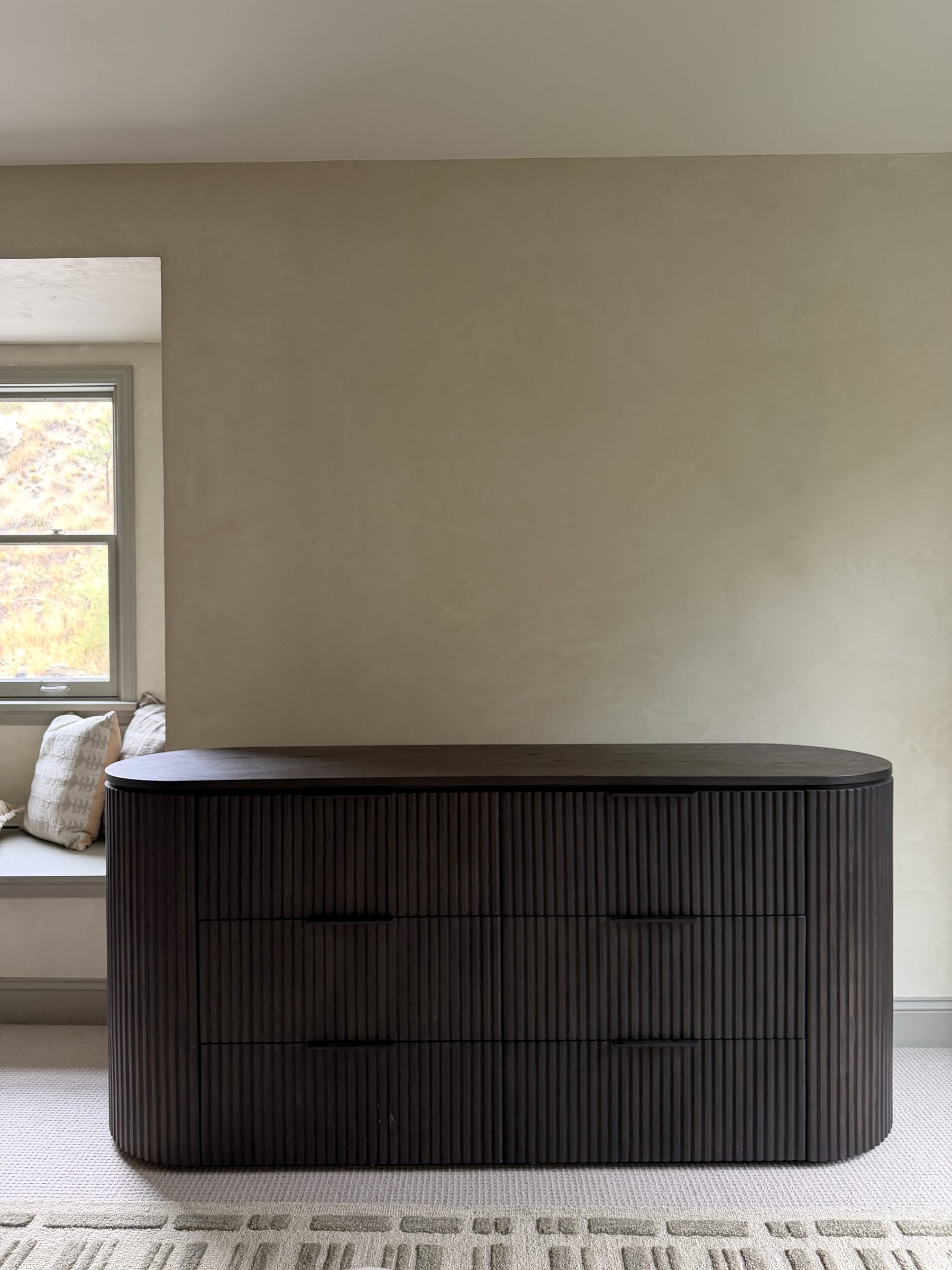 A dark fluted wood dresser with a completely bare top, set against an empty limewashed plaster wall beside a window seat