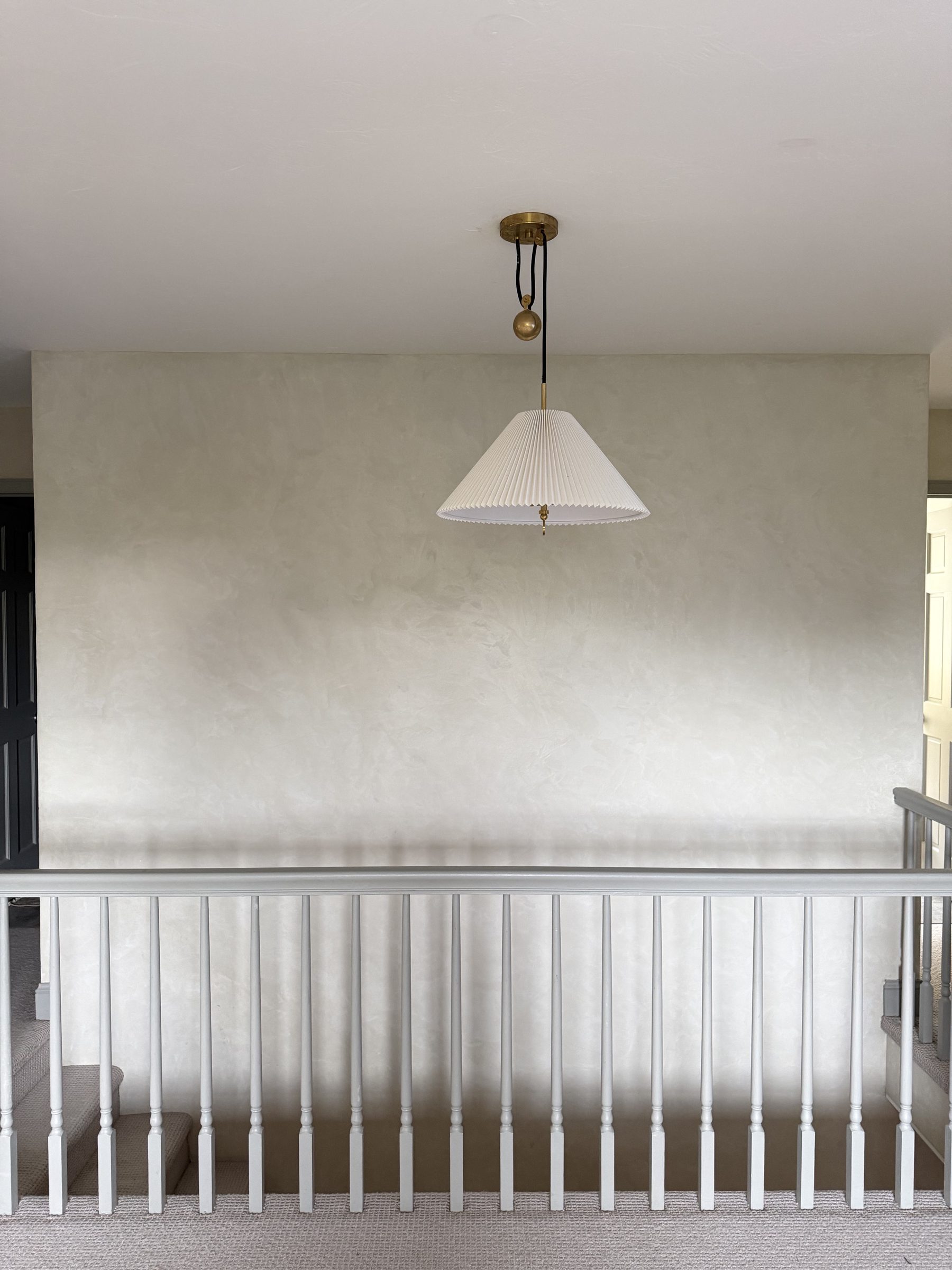 A large empty limewashed plaster wall at the top of a staircase, with a pleated pendant light and painted railing