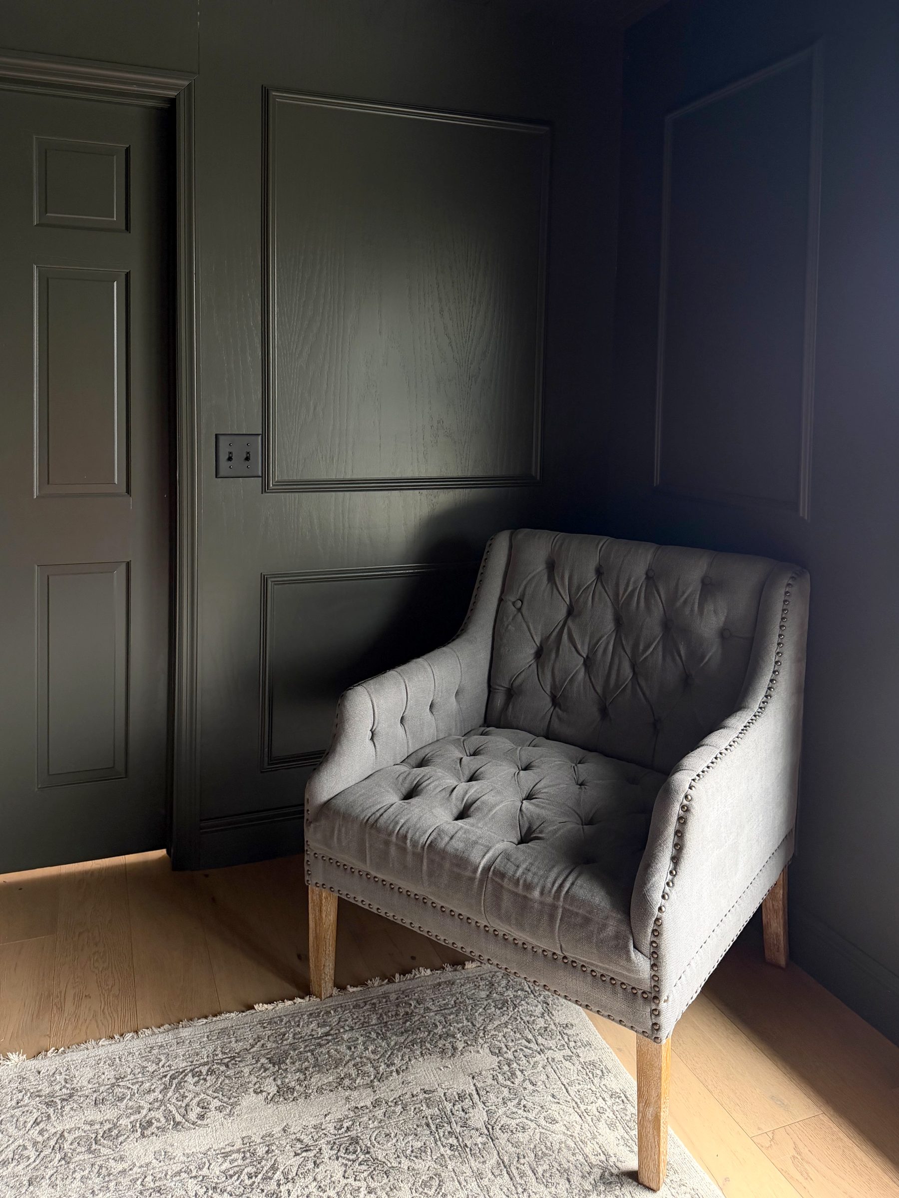 A corner painted deep green with panelled millwork, a grey tufted armchair, and white oak floors in low directional light
