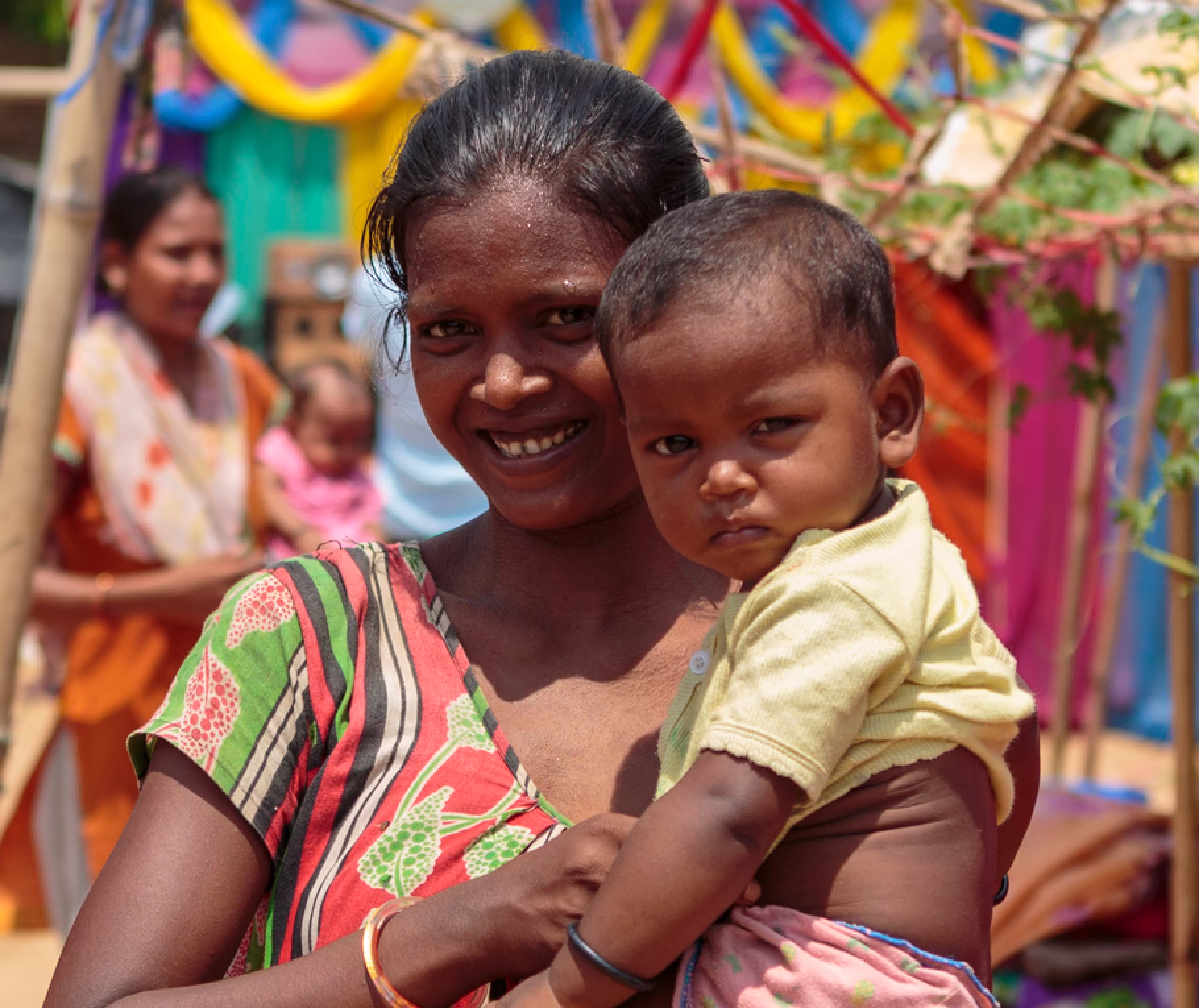 Smiling woman holding a young child with colorful fabric and people in the blurred background outdoors.