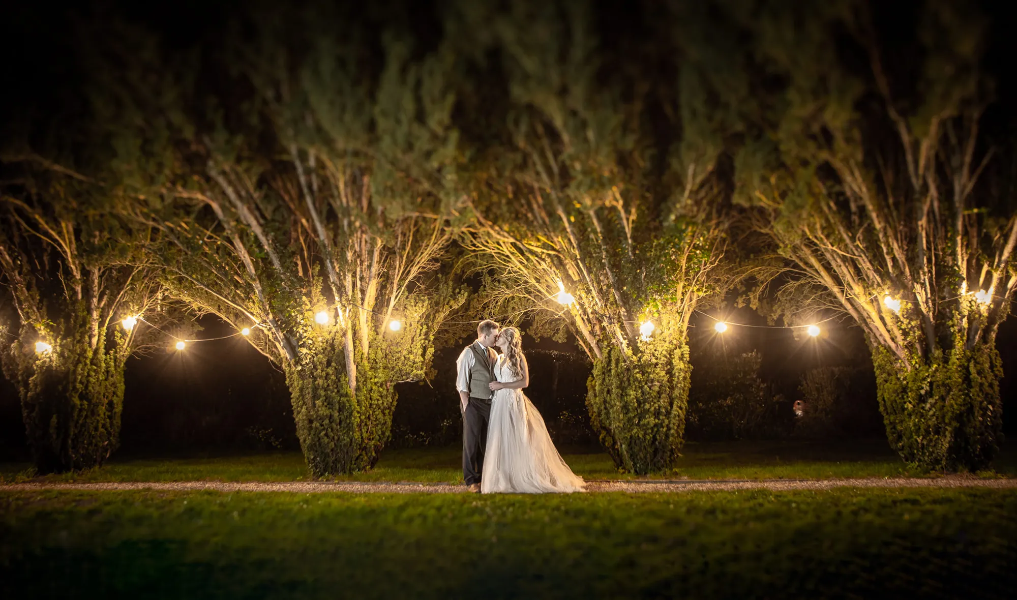 A bride and groom between yew trees lit by festoon lights at night at Lanwades Hall