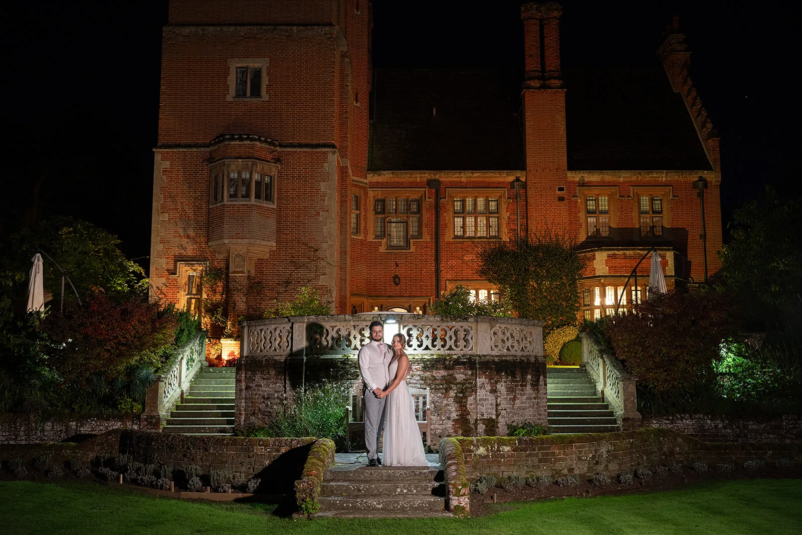 A bride and groom on the terrace steps in front of the lit house at night at Lanwades Hall