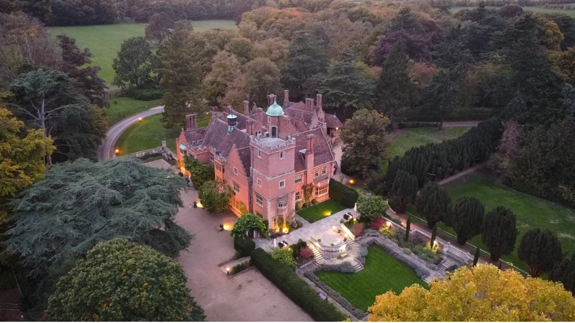 Aerial view of Lanwades Hall lit up at dusk, surrounded by gardens and woodland