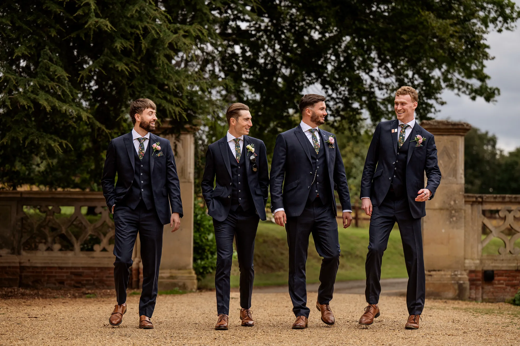 A groom and groomsmen in navy three-piece suits walking past the stone gate piers at Lanwades Hall