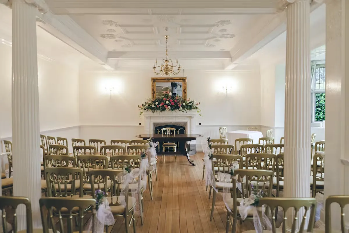 A bright pillared room with a chandelier set with gold chairs for a wedding ceremony at Lanwades Hall