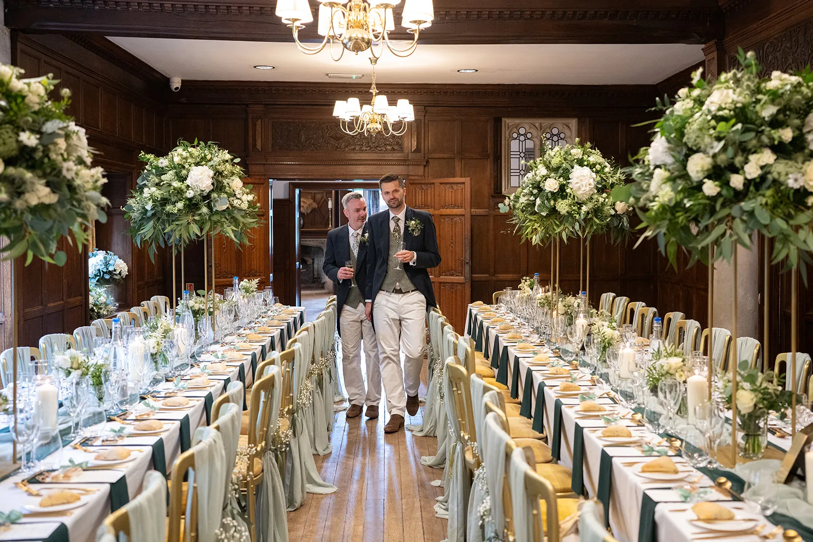 Two grooms walking between long flower-filled tables in the panelled hall at Lanwades Hall