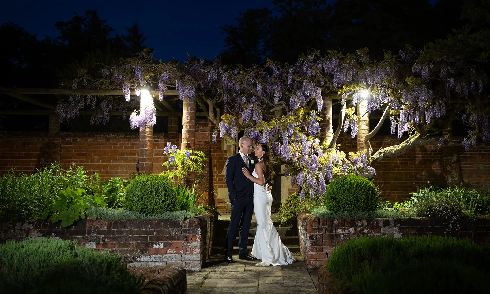 A bride and groom under lit wisteria in the walled garden at night at Lanwades Hall