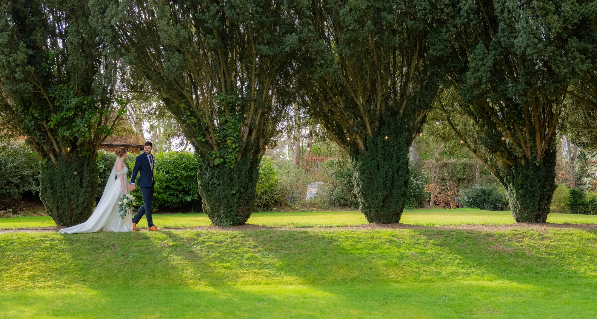 A bride and groom walking by the clipped yews in the garden at Lanwades Hall
