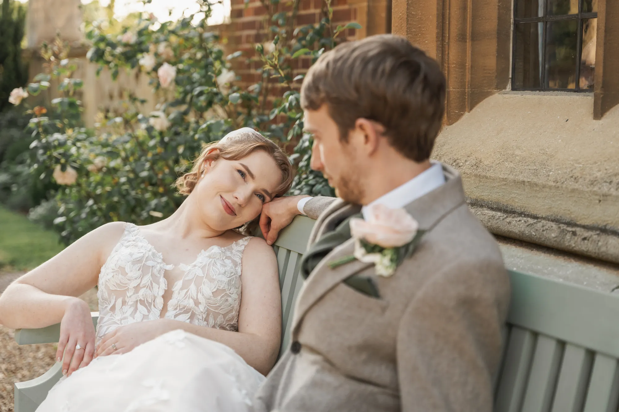 A bride resting her head on the groom's shoulder on a garden bench among roses at Lanwades Hall