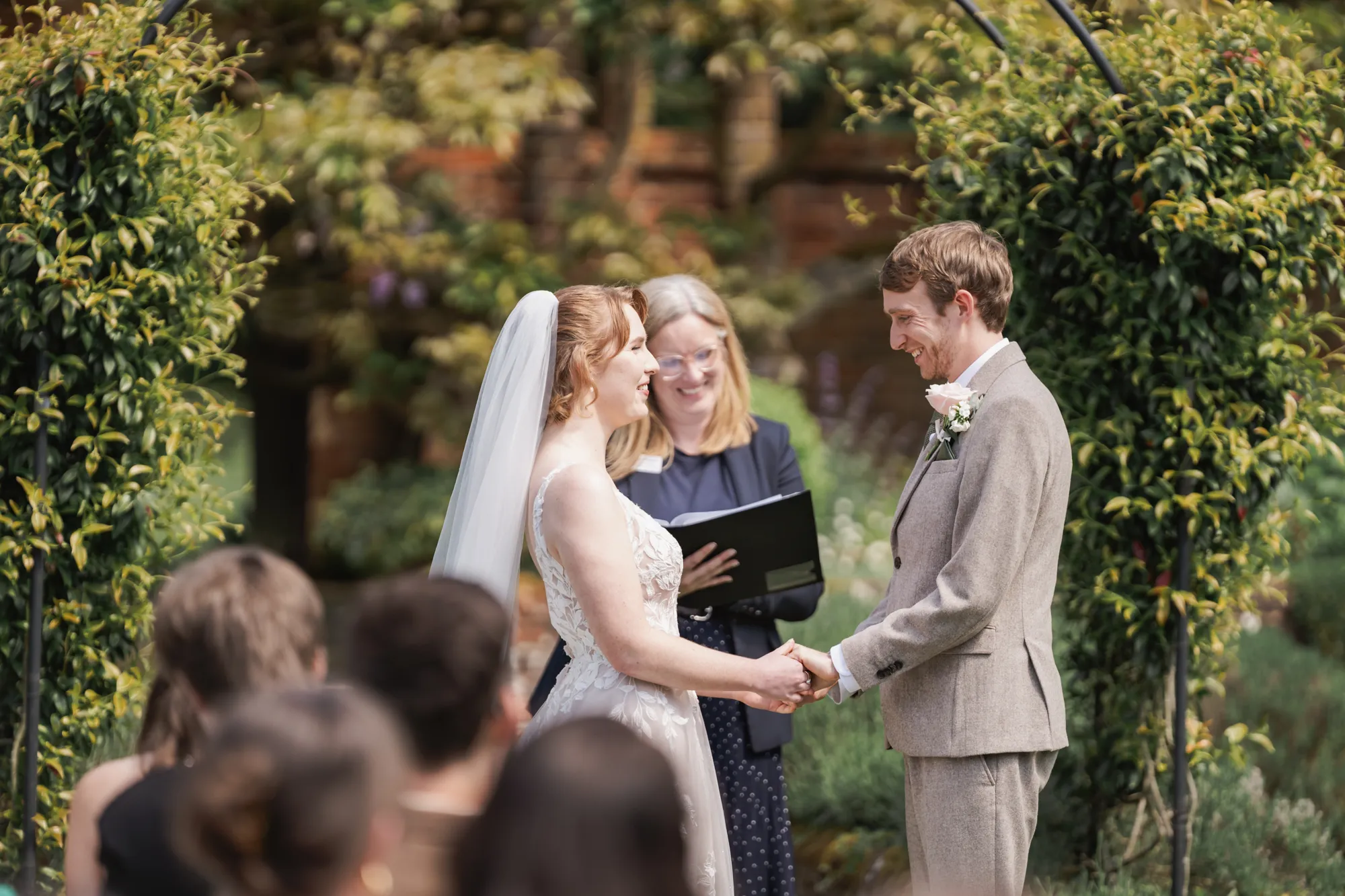 A bride and groom holding hands under a garden arch during their outdoor ceremony at Lanwades Hall