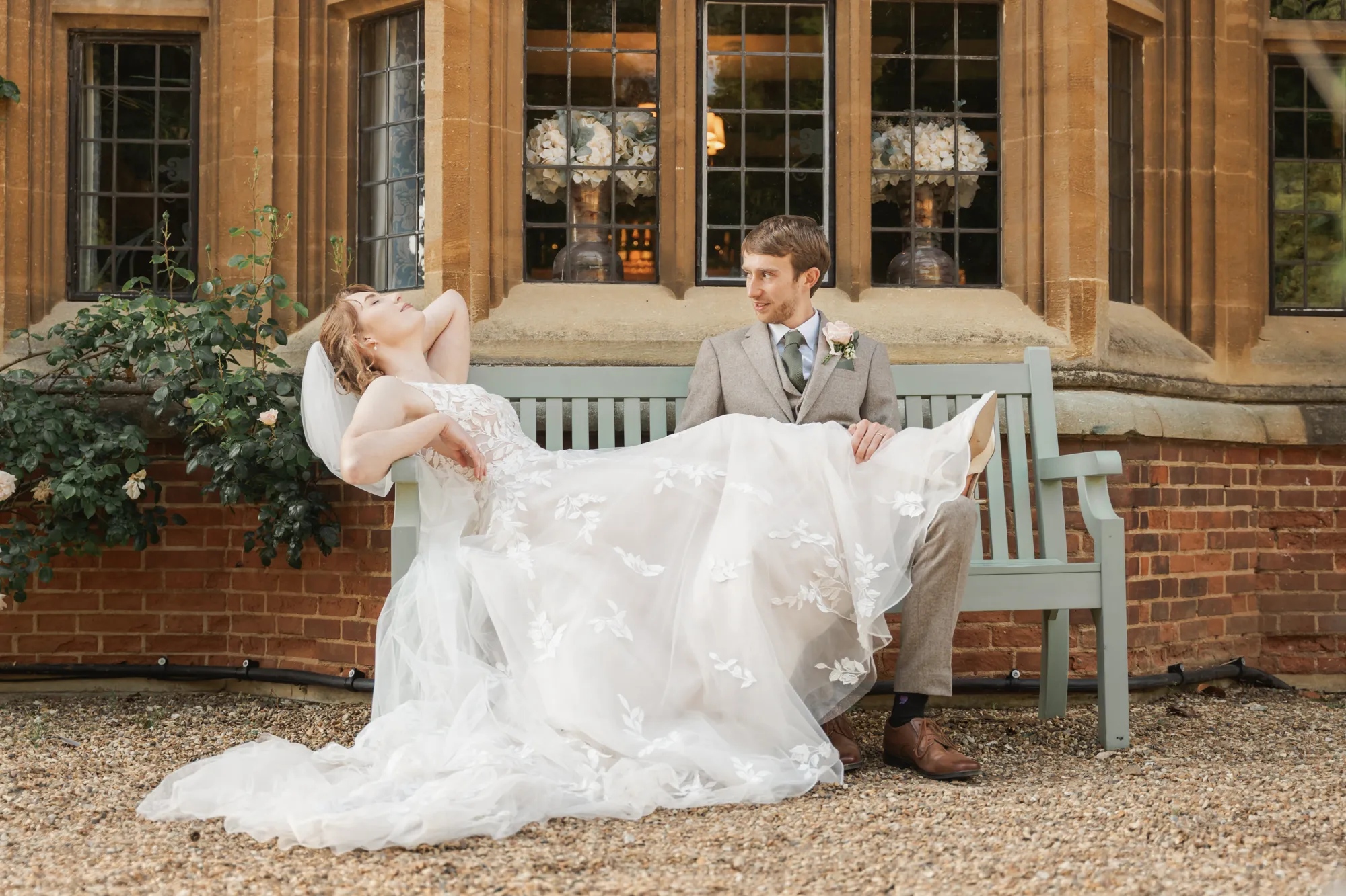 A relaxed bride and groom on a bench beneath the stone bay window of Lanwades Hall