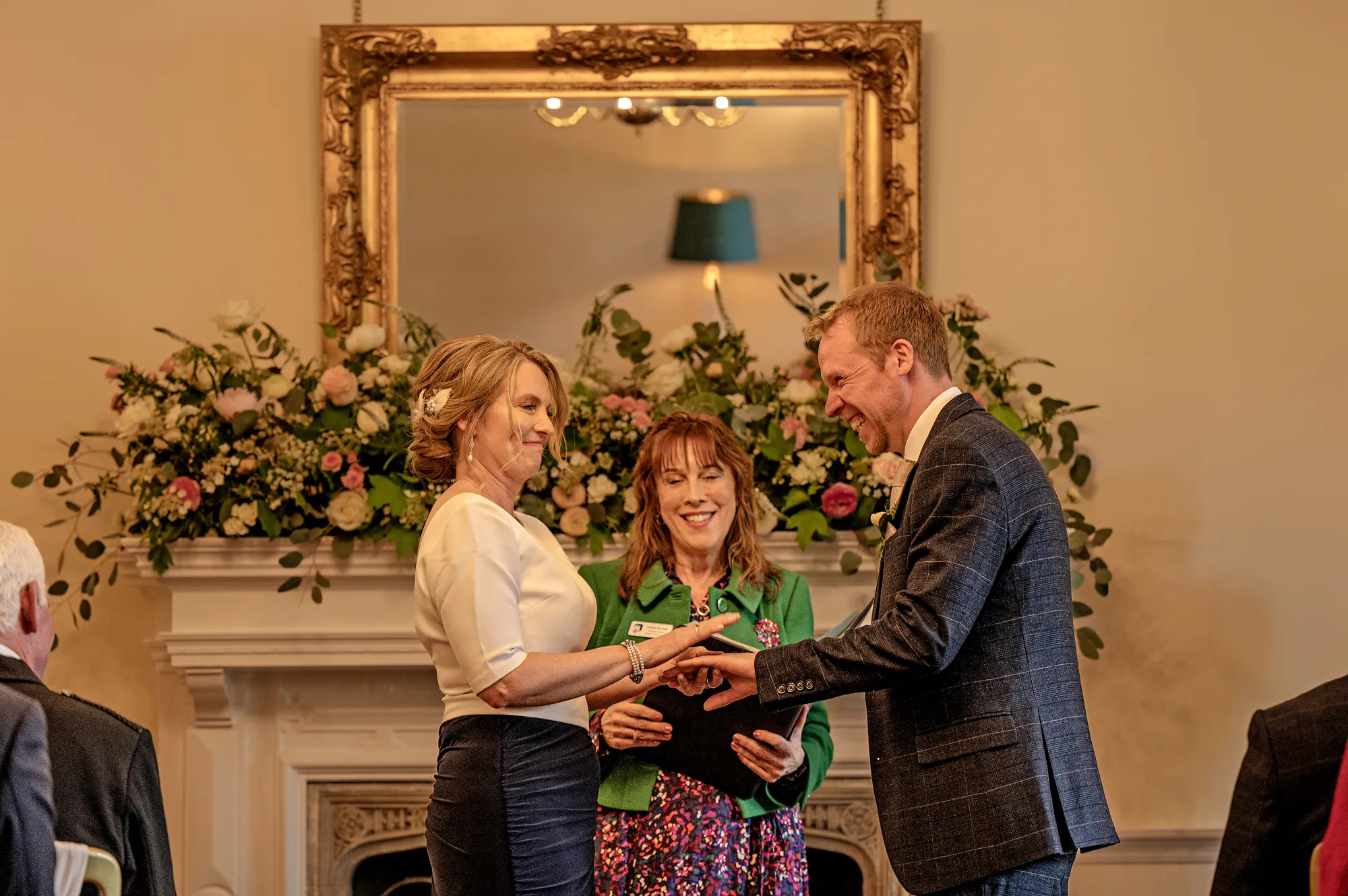 A couple exchanging rings by the flower-dressed fireplace and gilt mirror at Lanwades Hall