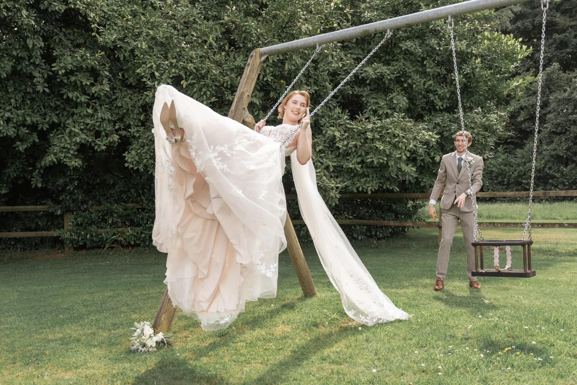 A bride on the garden swing with her dress flying as the groom watches at Lanwades Hall