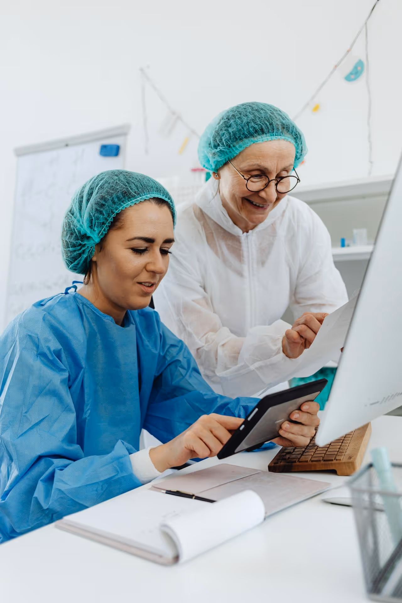 Two scientists in protective gear reviewing data on a tablet and paper in a lab.
