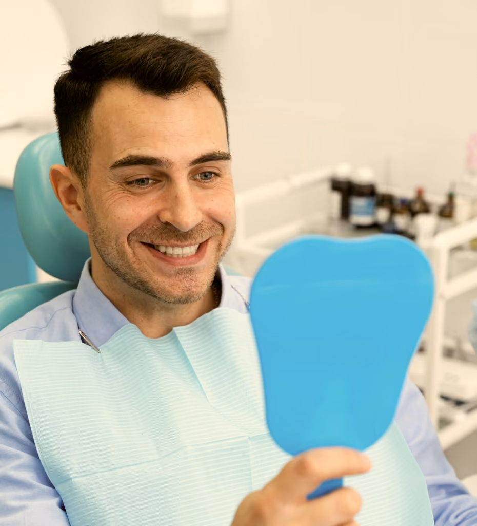 Smiling man sitting in a dental chair looking at his reflection in a blue tooth-shaped hand mirror, wearing a dental bib in a dental clinic.