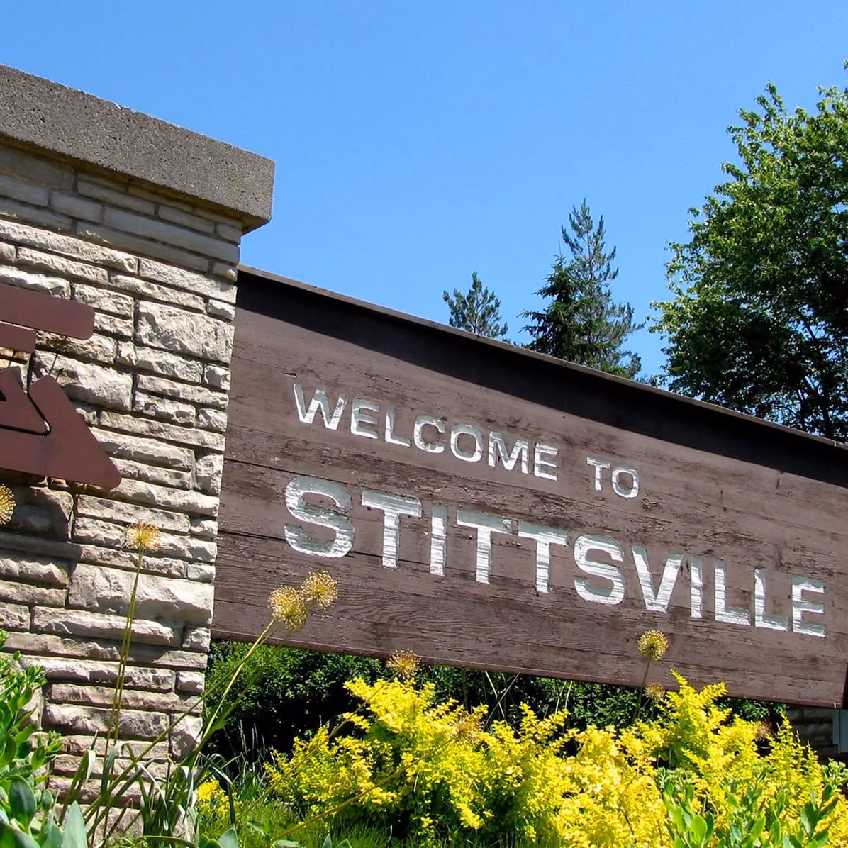 Wooden sign on a stone pillar with white letters reading 'WELCOME TO STITTSVILLE' surrounded by green shrubs and yellow flowers under a clear blue sky.