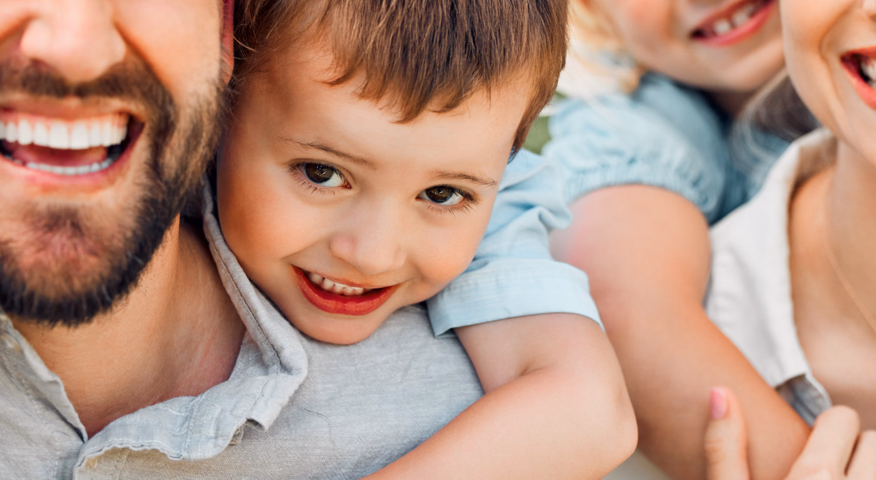 Close-up of a smiling child hugging a man from behind, with part of a woman and another child visible, all smiling.