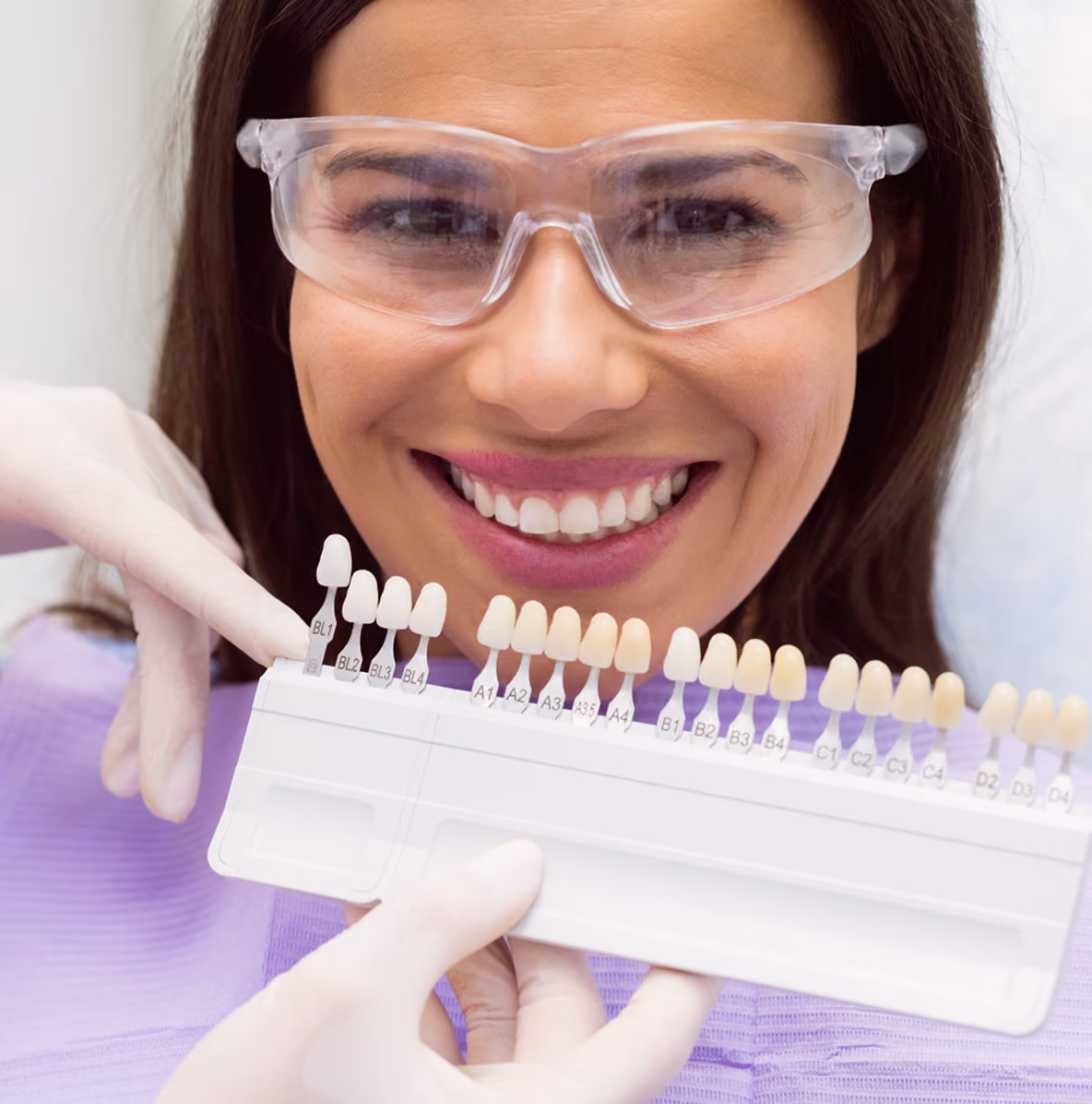 A smiling woman wearing protective glasses and a purple dental bib holds a shade guide with dental veneers or teeth color samples to match natural tooth color.