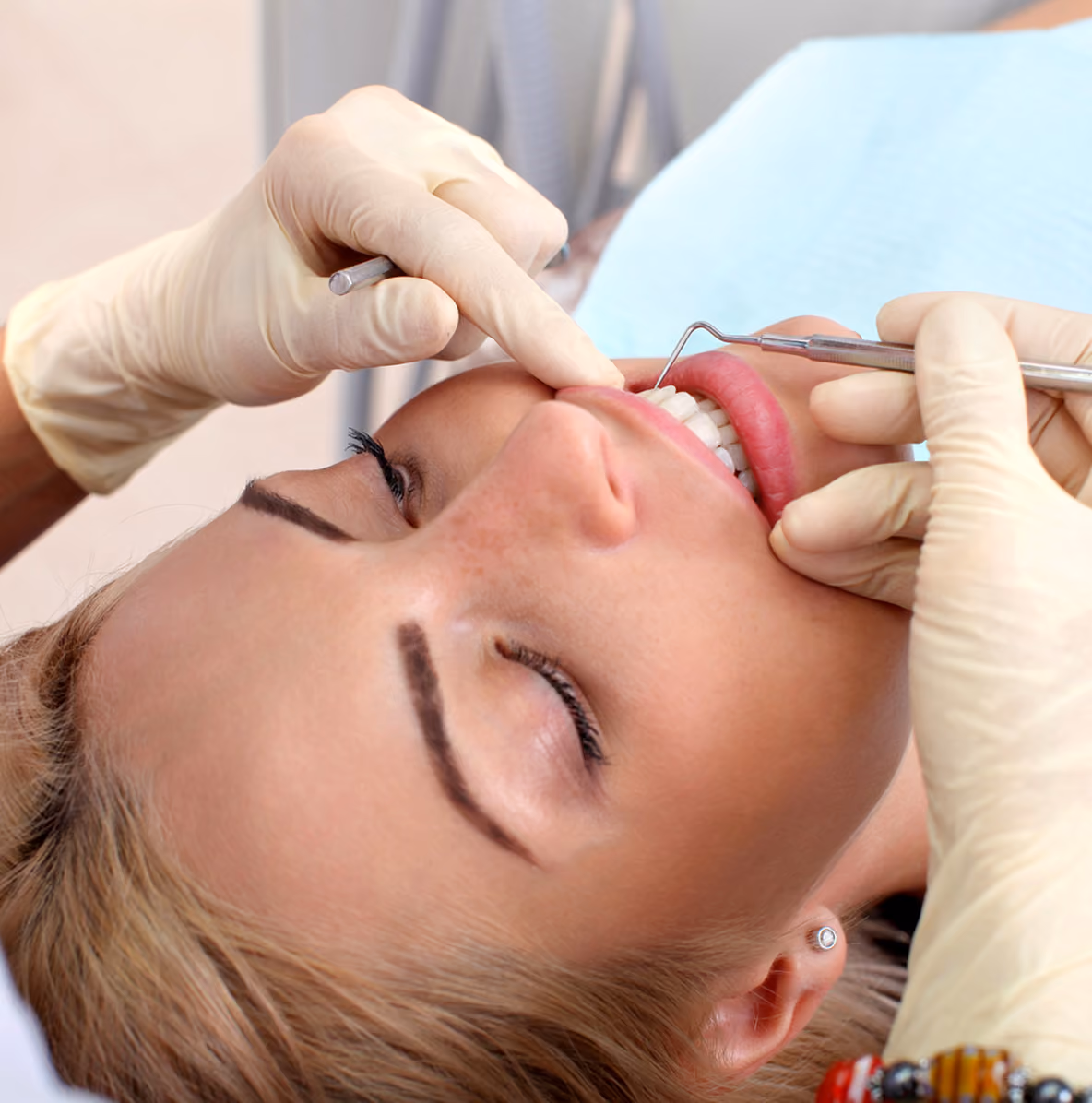 Close-up of a woman receiving dental care, with gloved hands holding dental tools examining her teeth as she lies back with eyes closed.