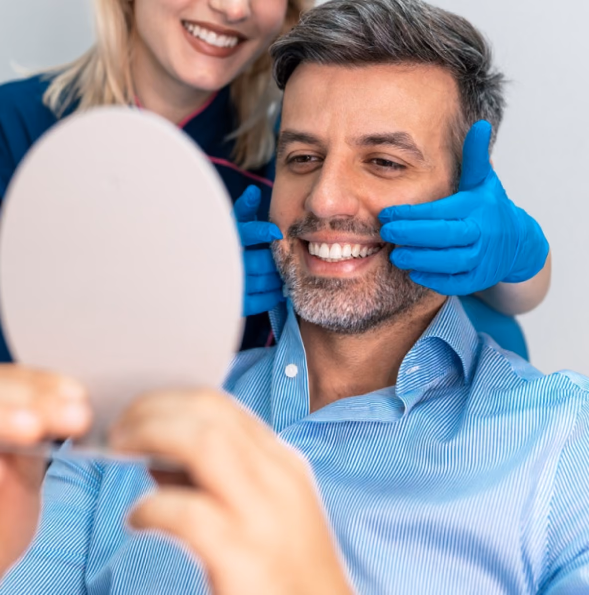 A man with salt-and-pepper hair and beard in a blue shirt smiles while looking into a hand mirror; a woman behind him wearing blue gloves gently holds his face, both appearing happy.