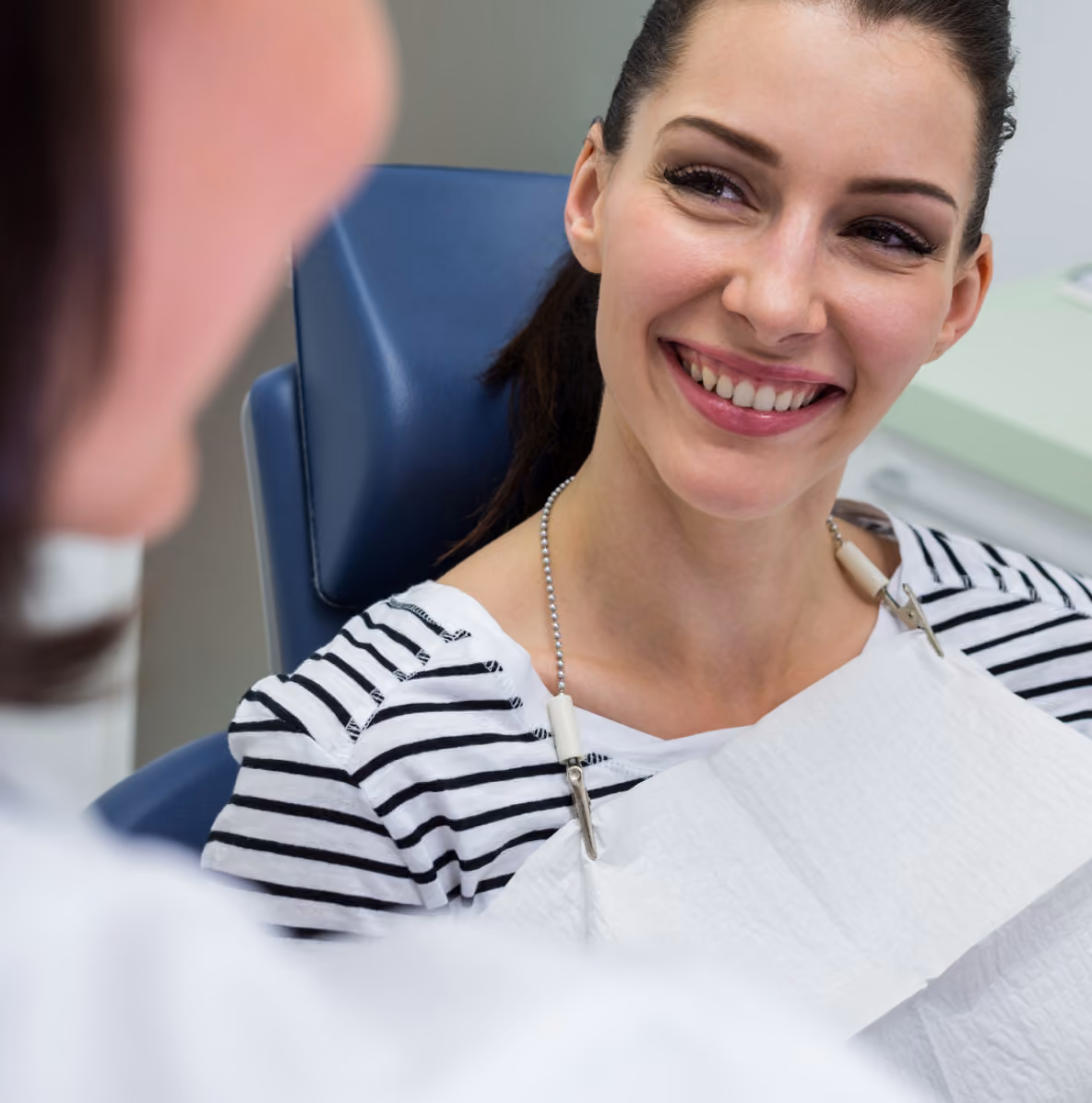 Woman in striped shirt sitting in a dental chair smiling at a person partially visible in the foreground.
