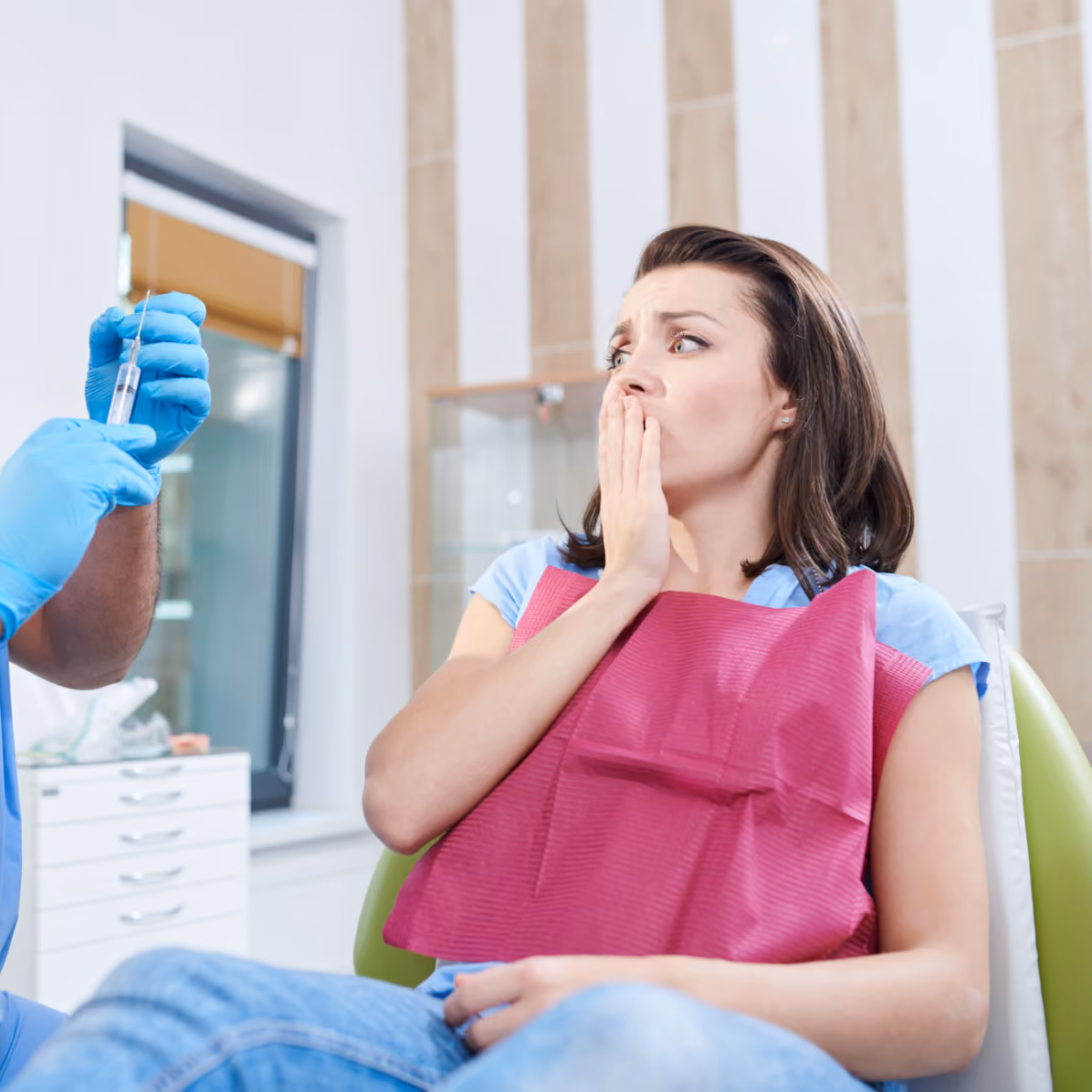 Woman sitting in a dentist chair looking anxious and covering her mouth with her hand while a healthcare professional wearing blue gloves holds a syringe.