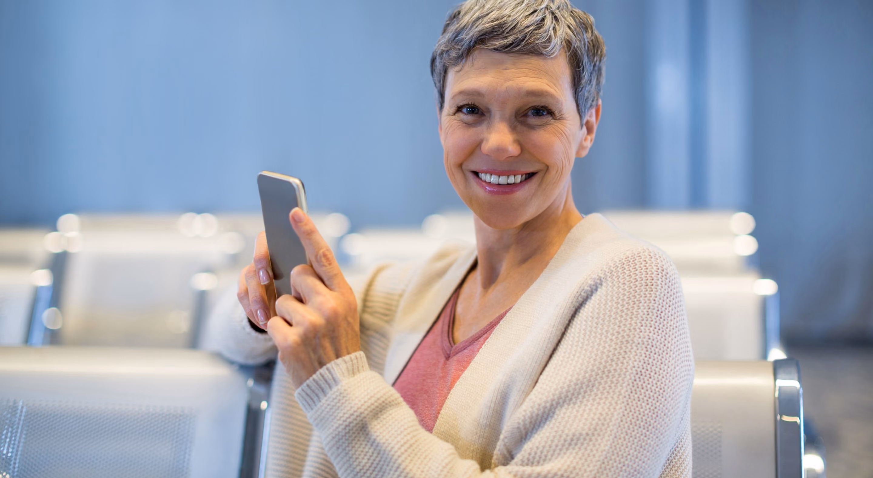 Smiling middle-aged woman with short gray hair wearing a white sweater and pink top, holding a smartphone, sitting on a metal chair in a waiting area.