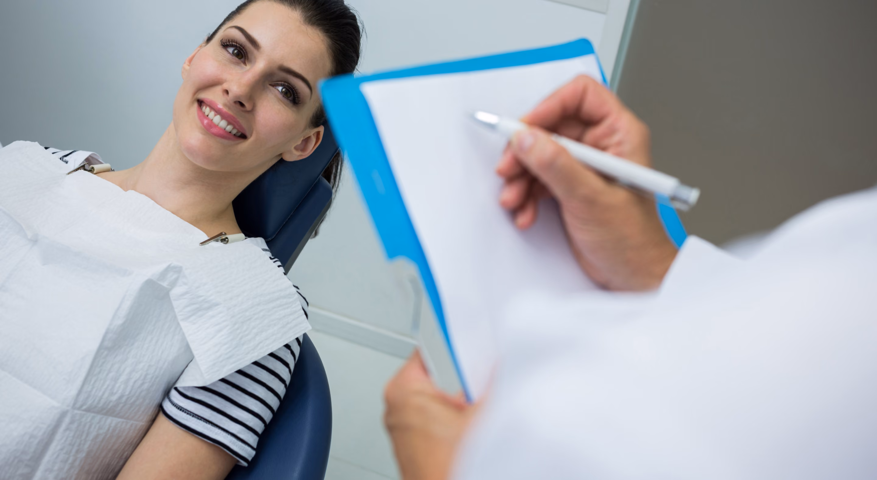 A smiling woman wearing a striped shirt reclines in a dental chair while a dentist holds a clipboard and pen, preparing to write.