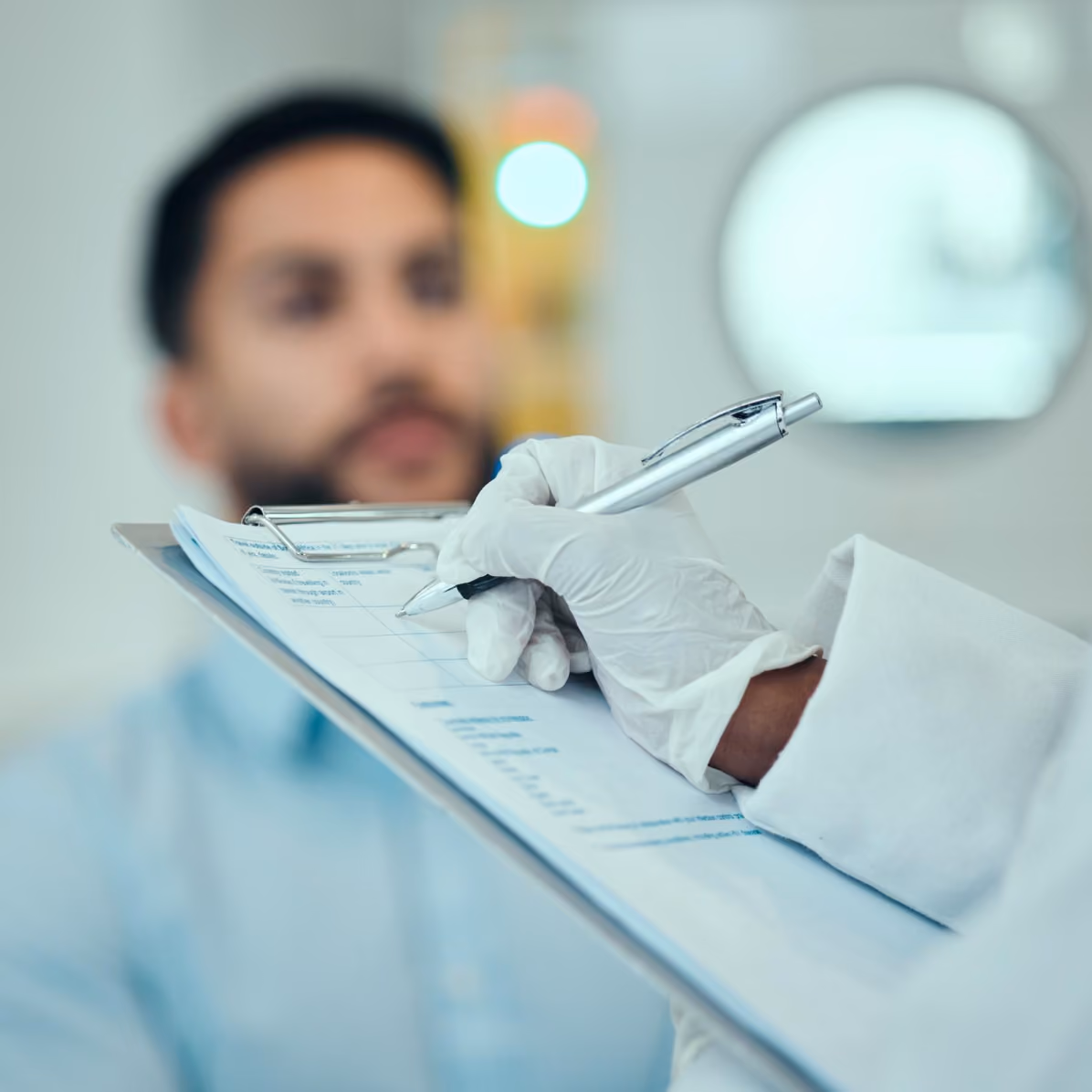 Medical professional wearing white gloves holding a pen and filling out a form on a clipboard with a blurred patient in the background.